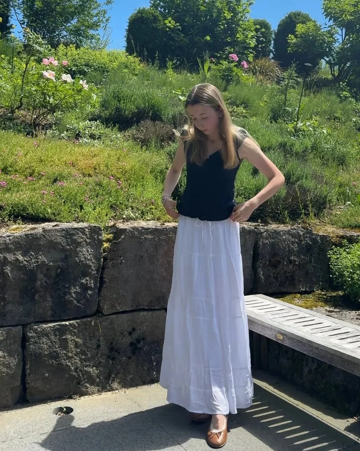 Junge Frau steht vor einem bepflanzten Steingarten, trägt ein dunkelblaues Top, einen langen weißen Rock und braune Ballerinas, bei strahlendem Sonnenschein.