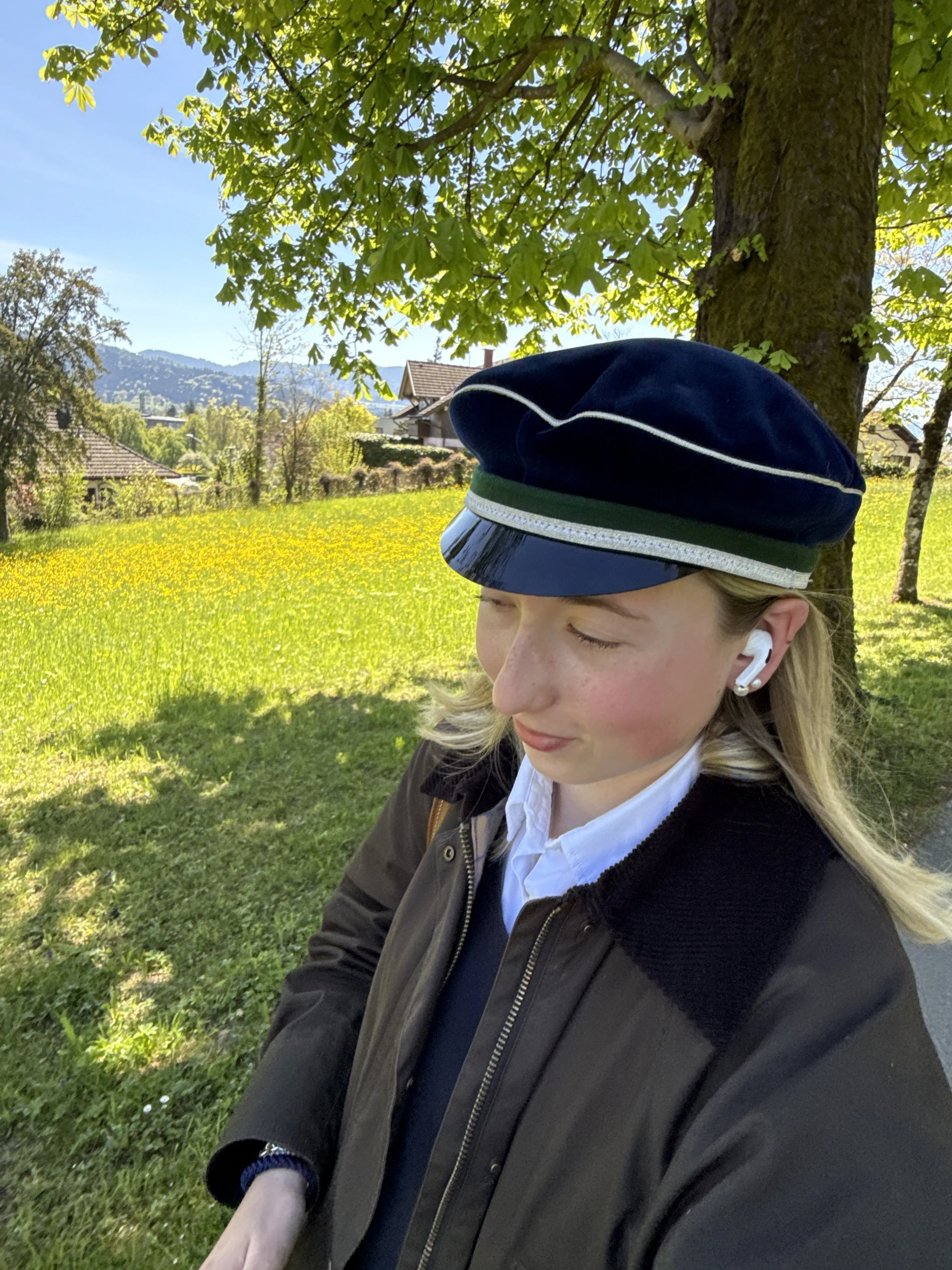 Ein Mädchen mit blonden Haaren, das eine blaue Mütze trägt, läuft im Freien unter einem Baum und trägt weiße AirPods, während sie entspannt und glücklich mit geschlossenen Augen ist.