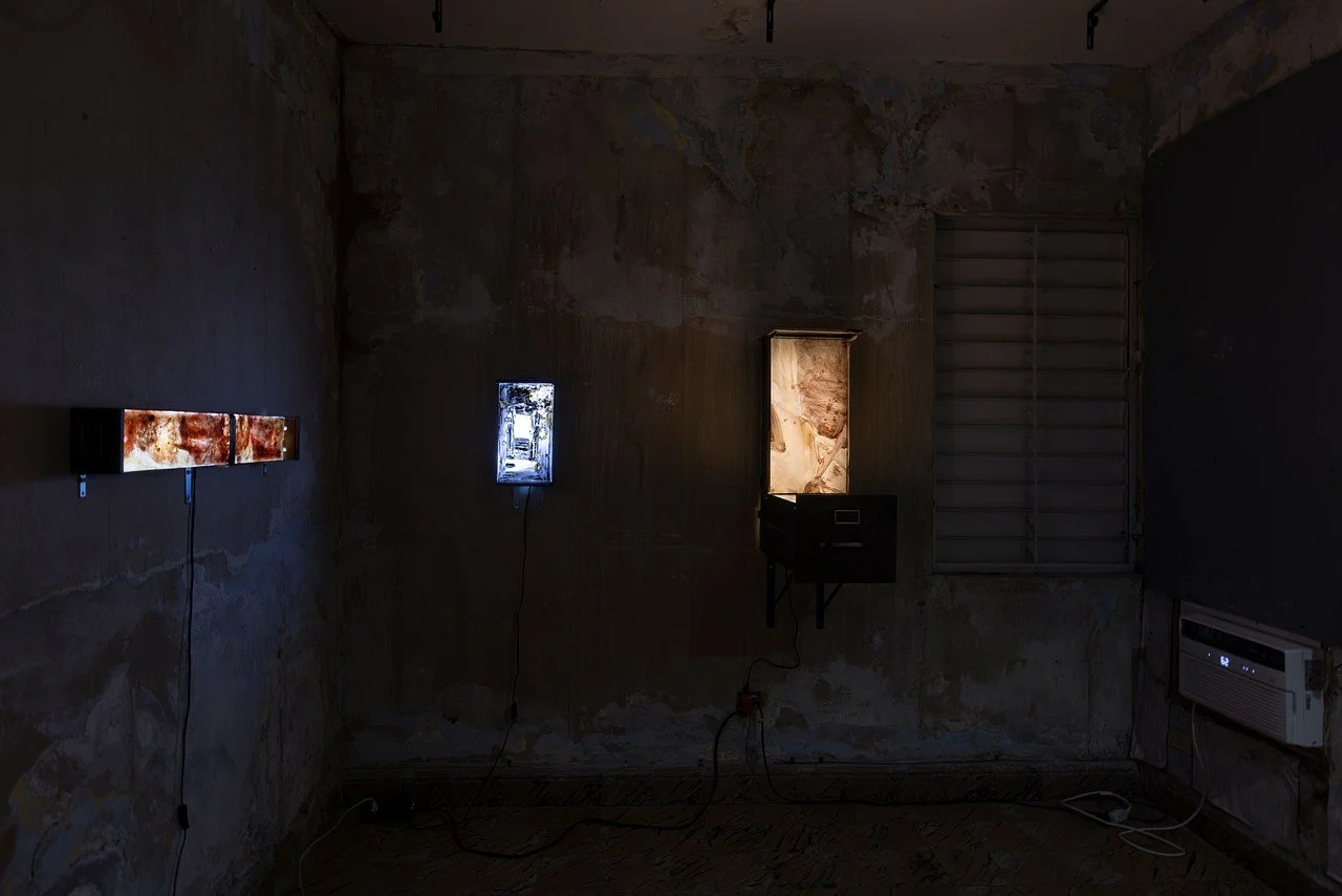 Dimly lit art installation with illuminated office cabinets, each displaying a painting and mounted on a textured wall in an empty room.