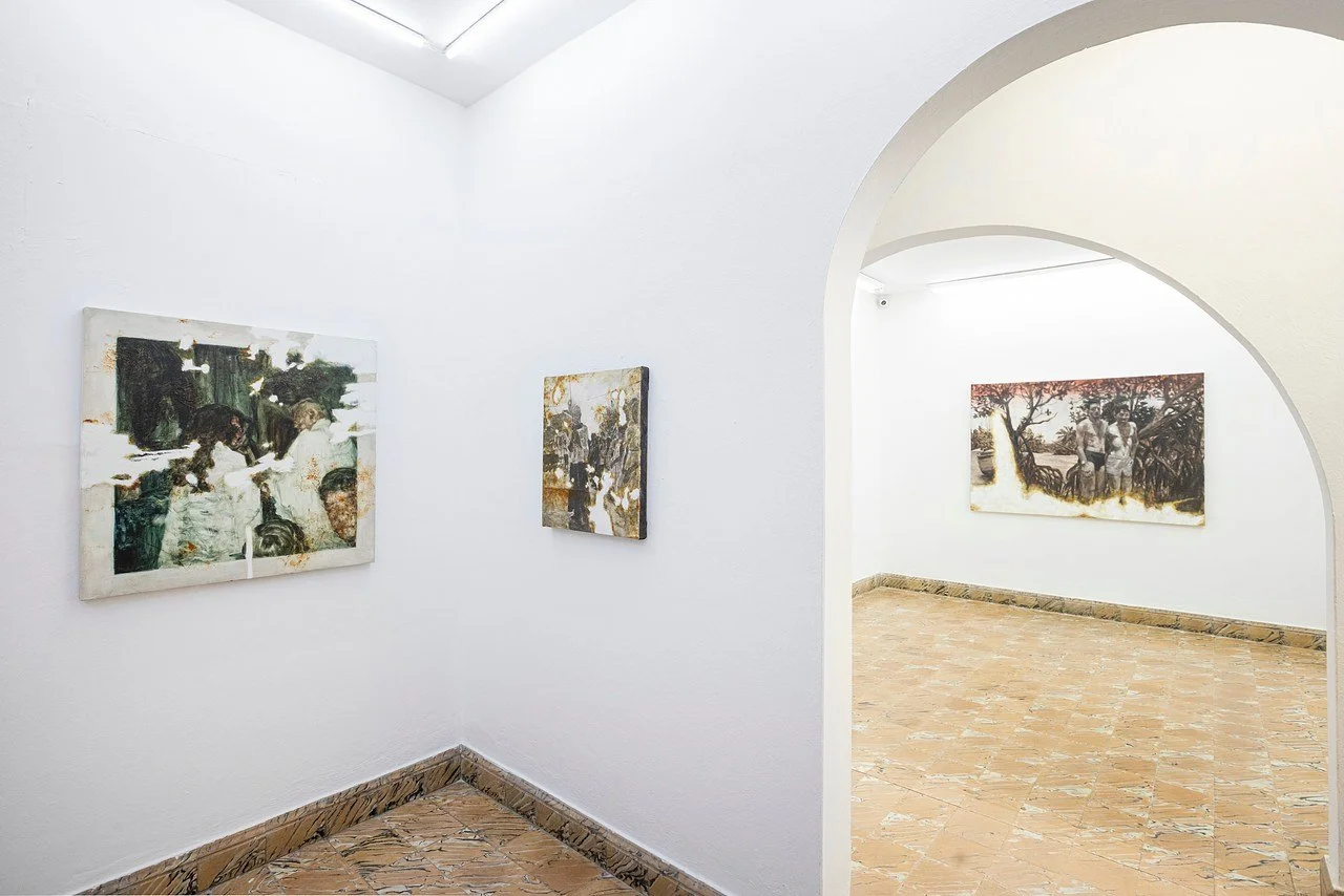An art gallery with white walls, displaying three paintings. One painting features two children, another depicts a group of people, and the third shows two people under a mangrove.