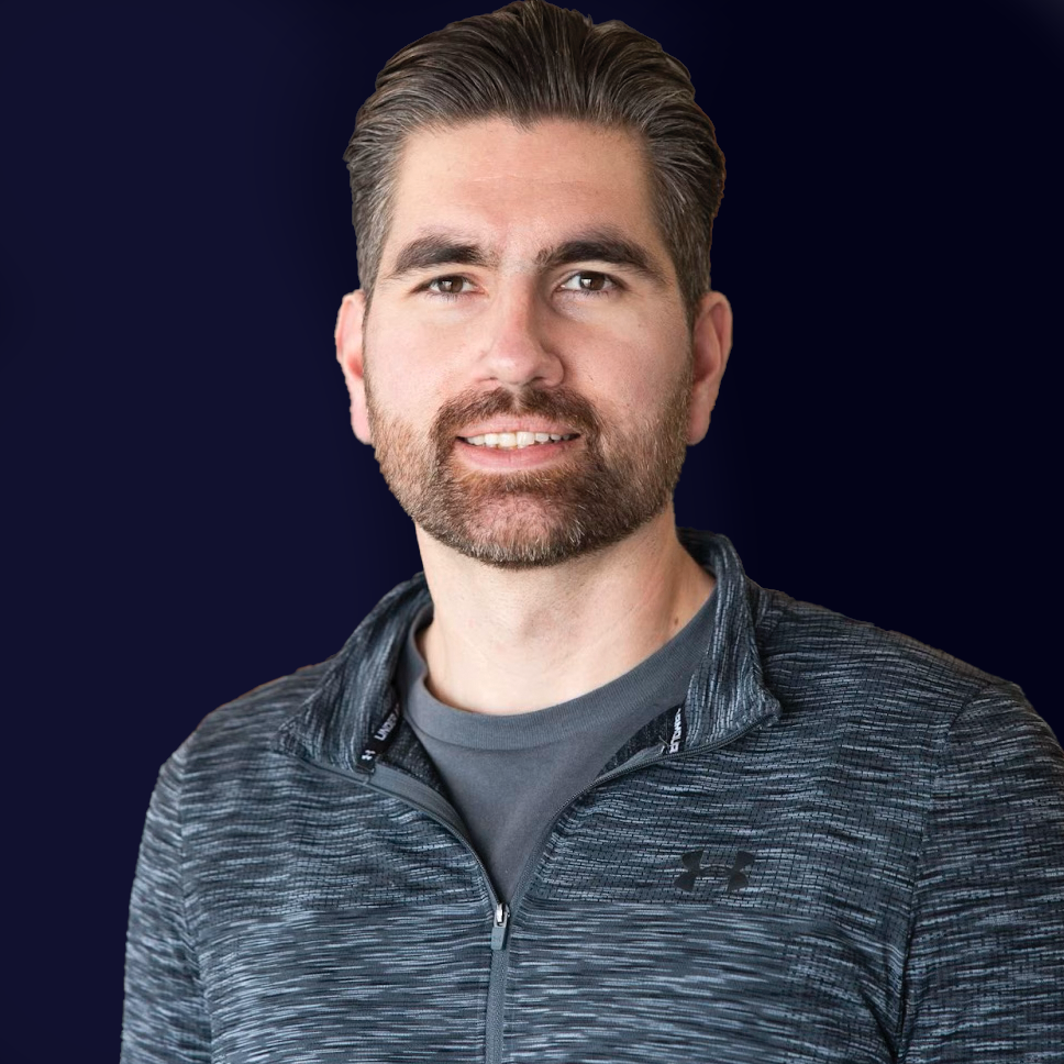 Portrait of a man with short brown hair, a beard, wearing a gray Under Armour zip-up athletic jacket, smiling slightly, against a dark background.