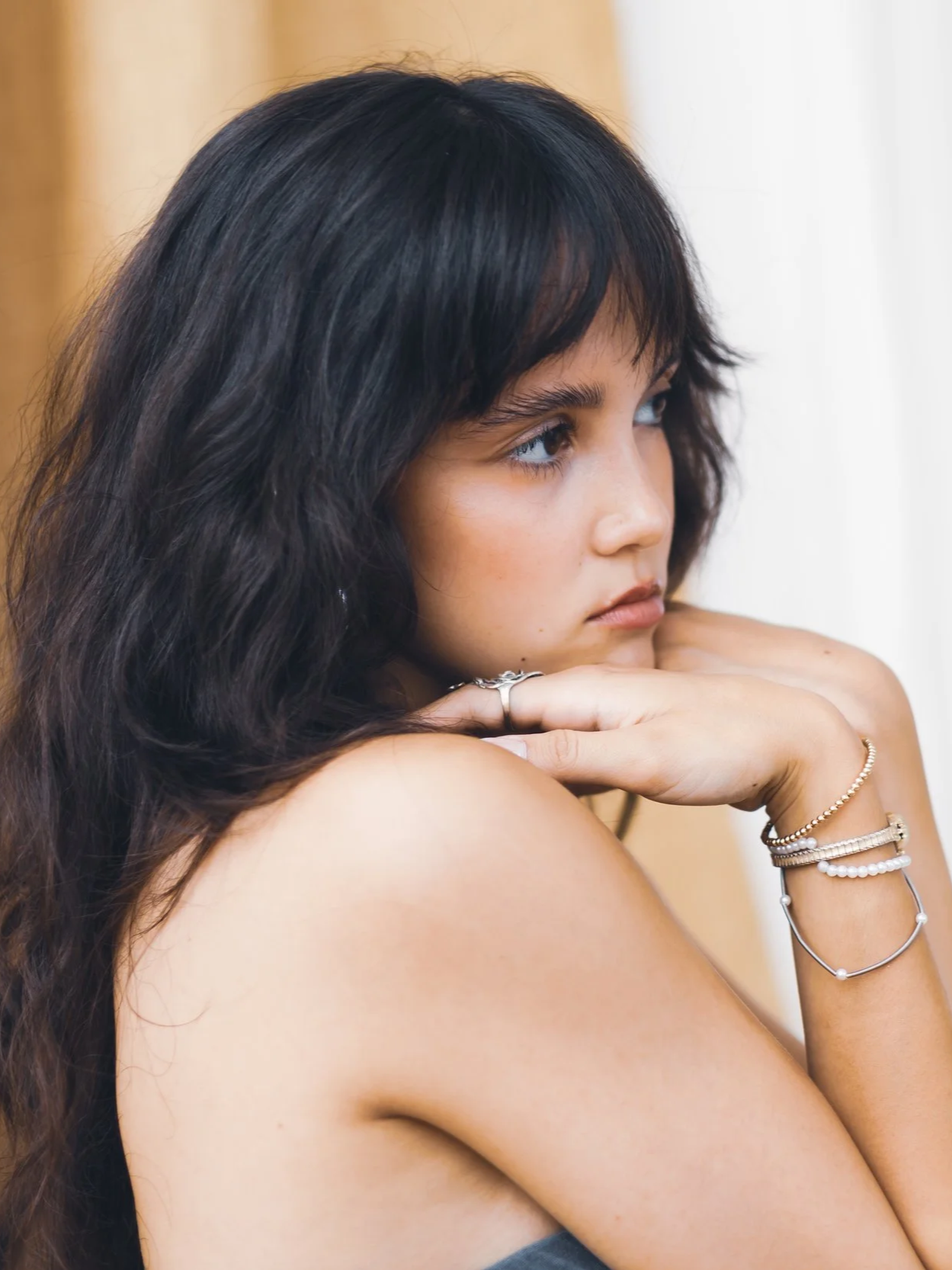 A young woman with long dark hair resting her chin on her arms, looking to the side with a contemplative expression, wearing bracelets and rings.