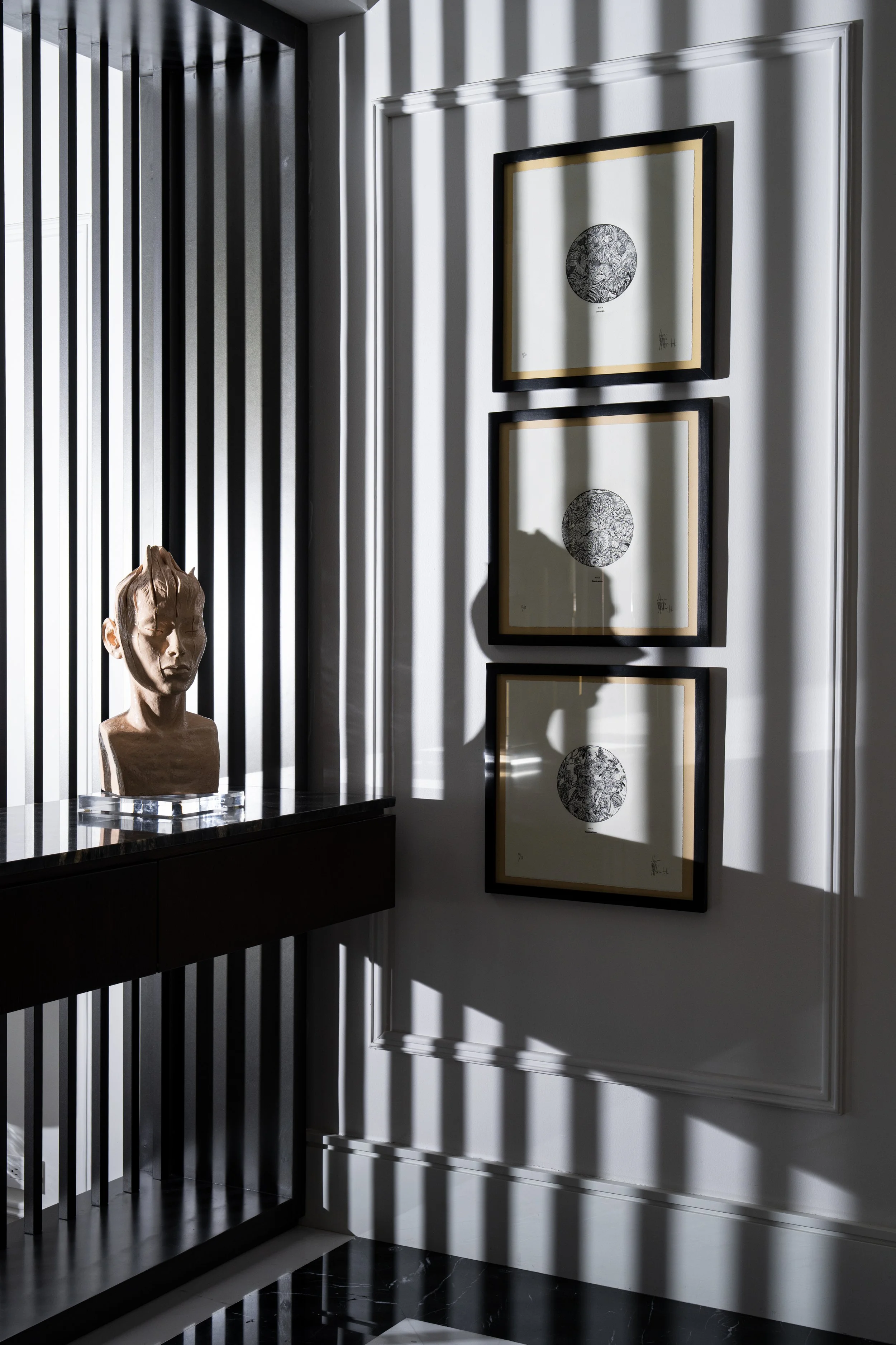 Entry foyer featuring a custom black marble floating console with a sculptural bust and three framed black-and-white botanical artworks. Dramatic natural light casts linear shadows across the paneled wall, creating a refined contemporary interior.