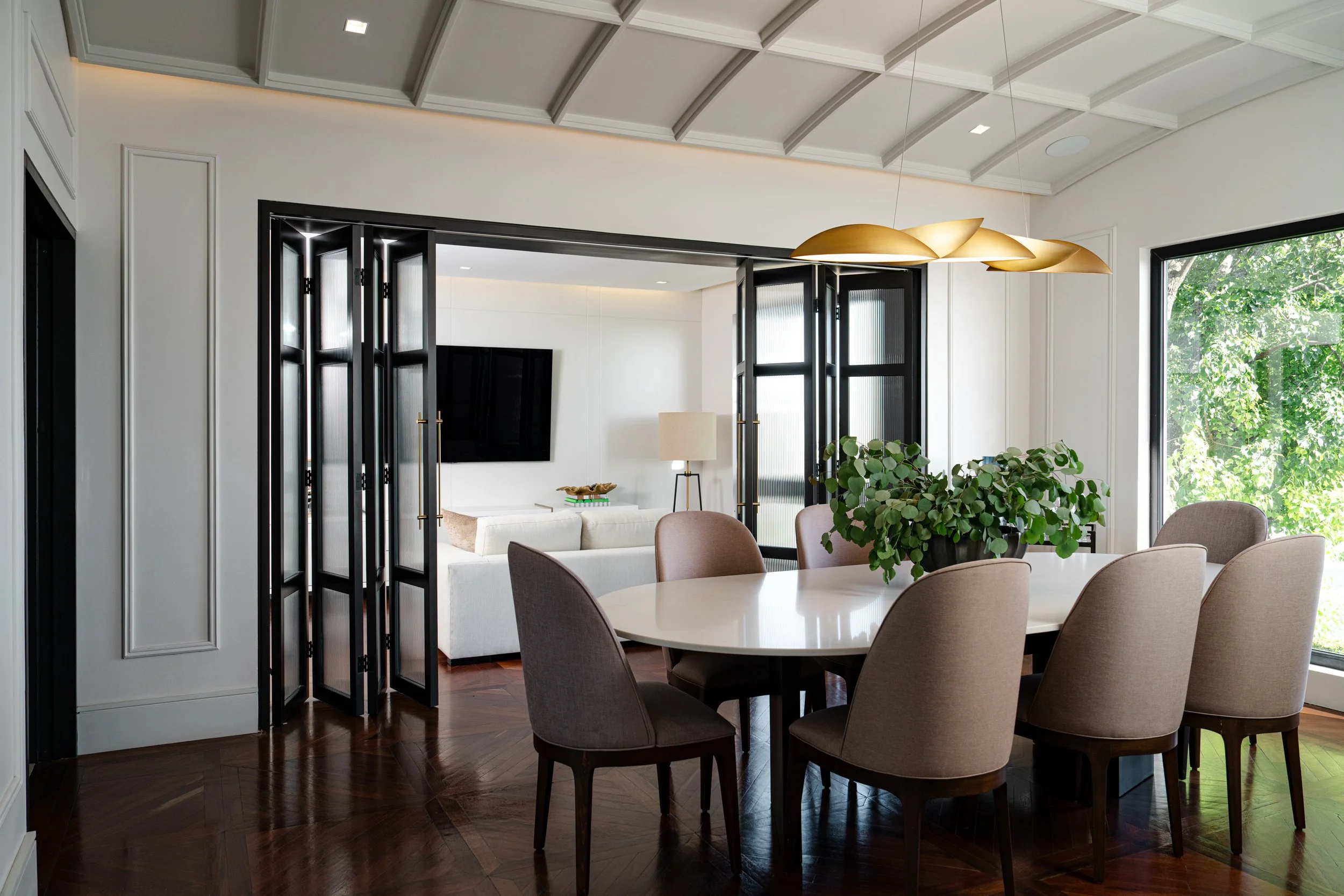 Modern dining room with a round table, upholstered chairs, large windows with greenery outside, and a decorative gold pendant light fixture.