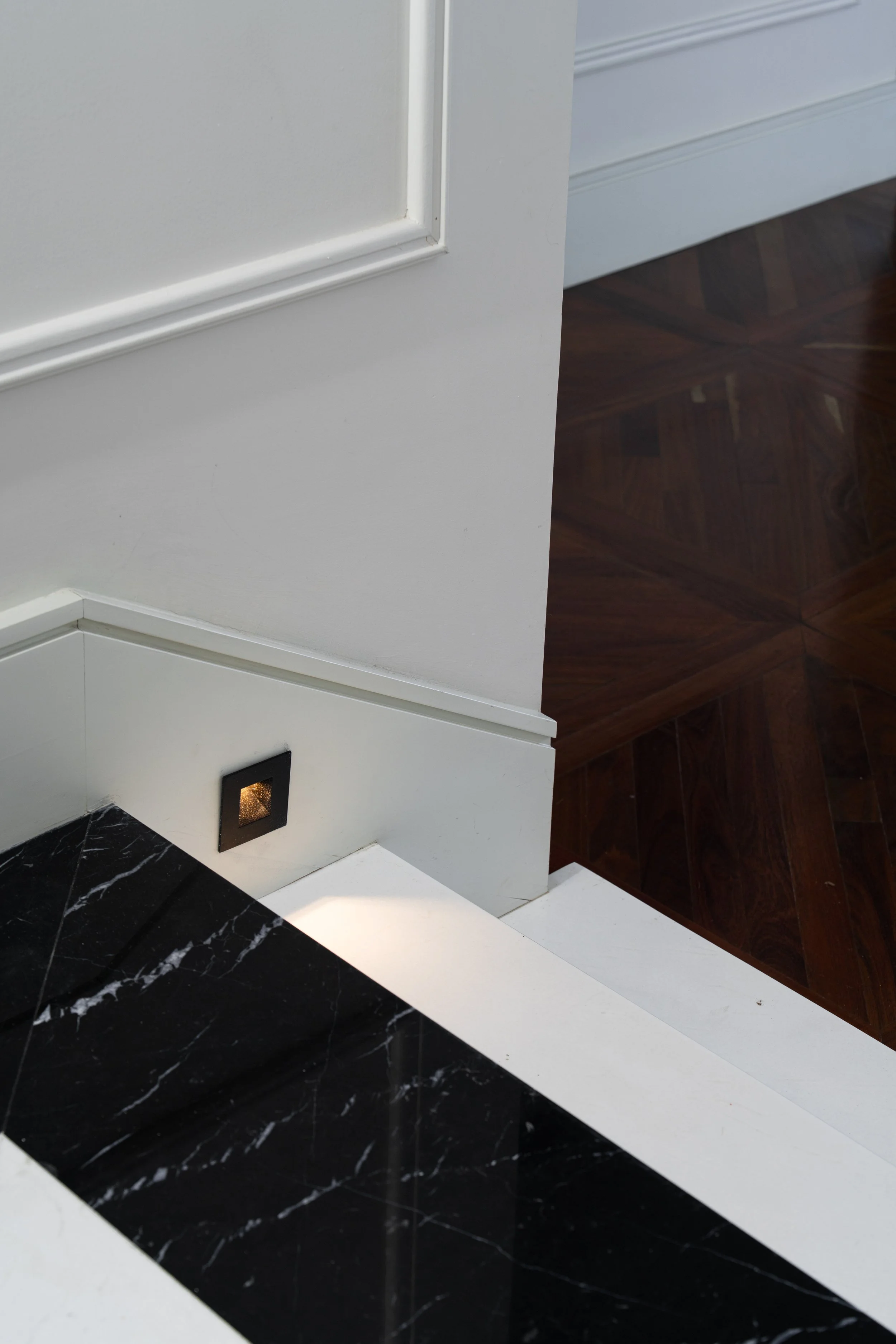 Contemporary staircase detail with black marble treads, white wall paneling, recessed step lighting, and dark wood parquet flooring, highlighting the contrast of natural materials.