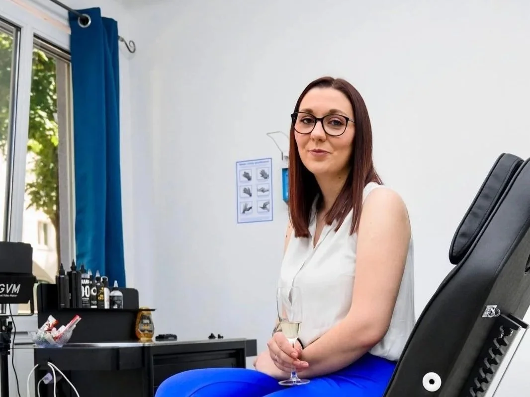 Eine Frau mit Brille sitzt auf einem Stuhl im Behandlungsraum eines Friseursalons, hält ein Glas Sekt in der Hand, neben ihr sind Haarpflegeprodukte sichtbar.
