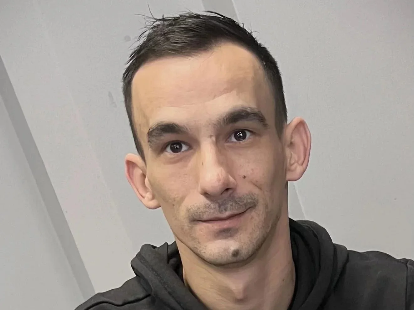 Nahaufnahme eines Mannes mit dunklen Haaren, braunen Augen und hellhäutiger Haut, trägt einen schwarzen Hoodie vor einem neutralen Hintergrund.
