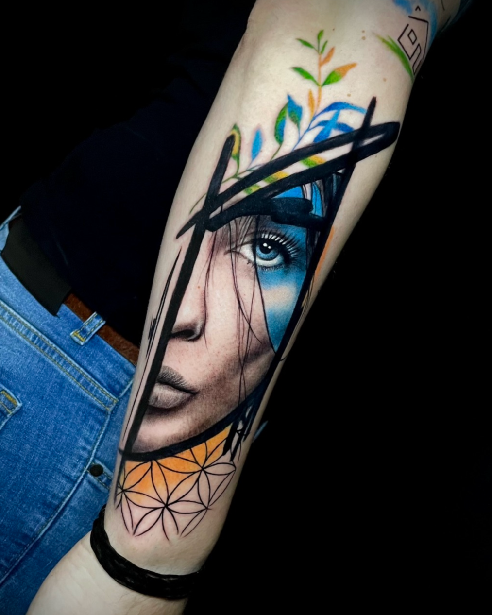 Tattoo eines menschlichen Gesichts mit einem blauen Auge, umgeben von geometrischen und naturinspirierten Mustern auf dem Arm.