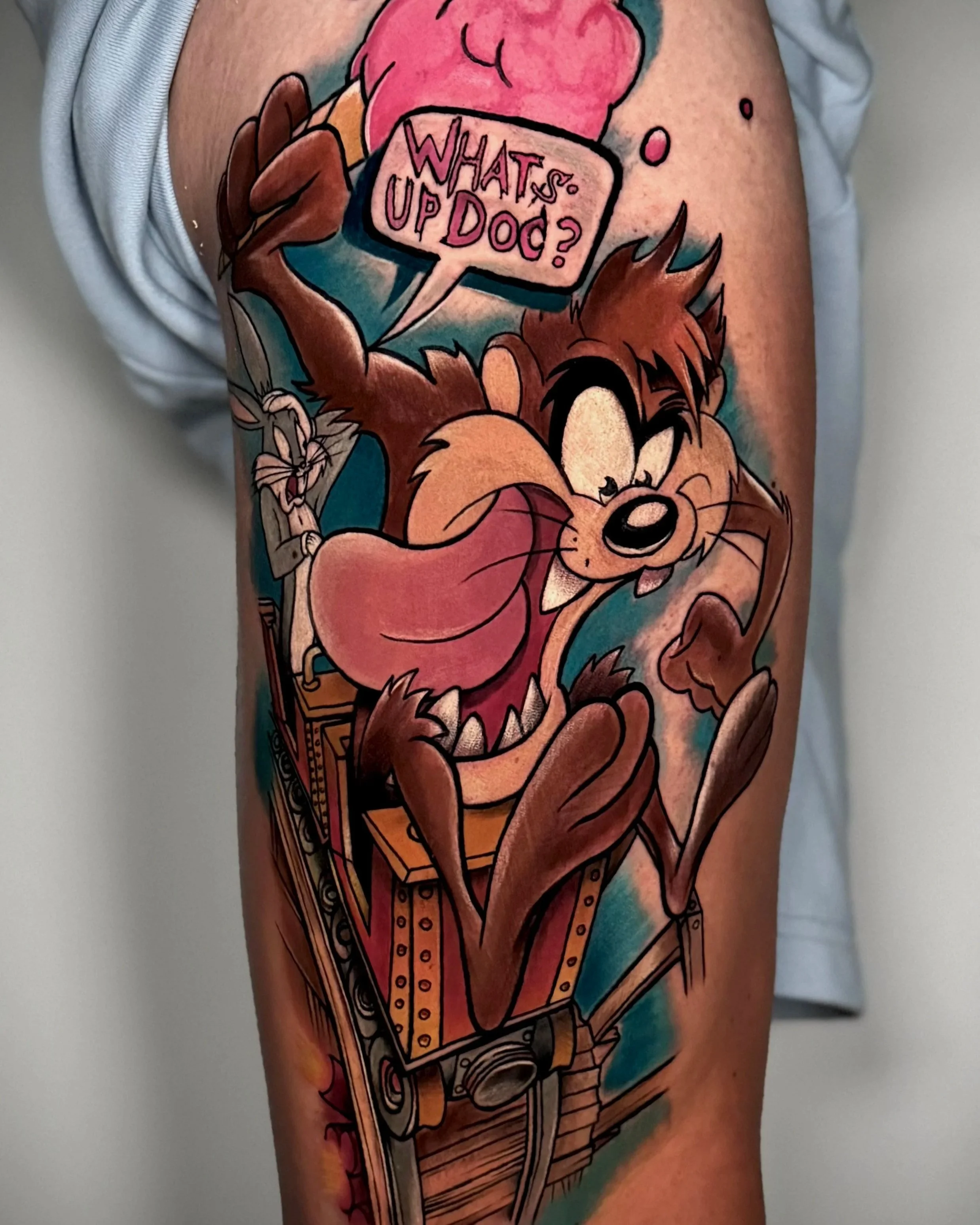 Tattoo auf dem Oberarm eines Menschen, das eine cartoonartige Figur eines knuddeligen Tigern mit einer Kamera zeigt, die eine Sprechblase mit dem Text 'WHAT'S UP DOC?' hält.