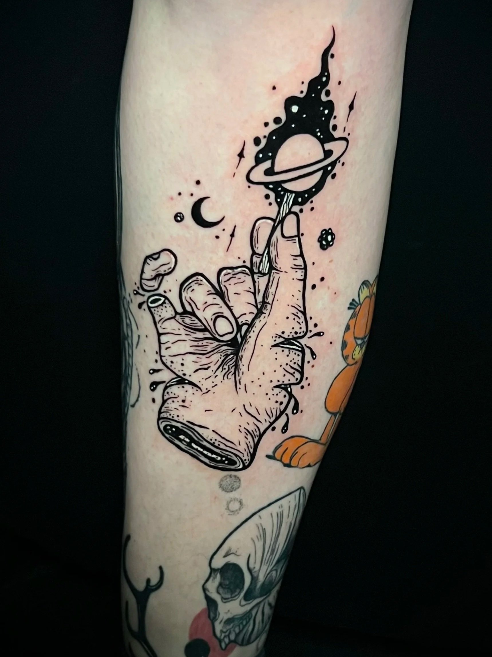 Tattoo auf Unterarm mit einem stilisierten Glas, einem Planet mit Ringen, einer Hand, einem Mond, kleinen Sternen und Punkten.