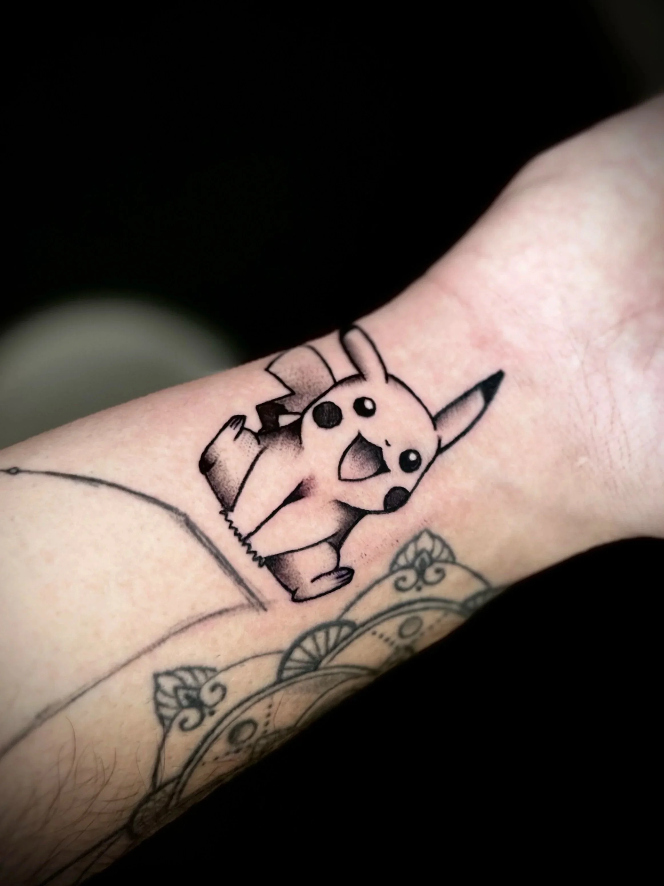 Tattoo eines Pikachu auf einem Unterarm, schwarz-weiße Skizze, mit Verzierung und Details im Hintergrund.