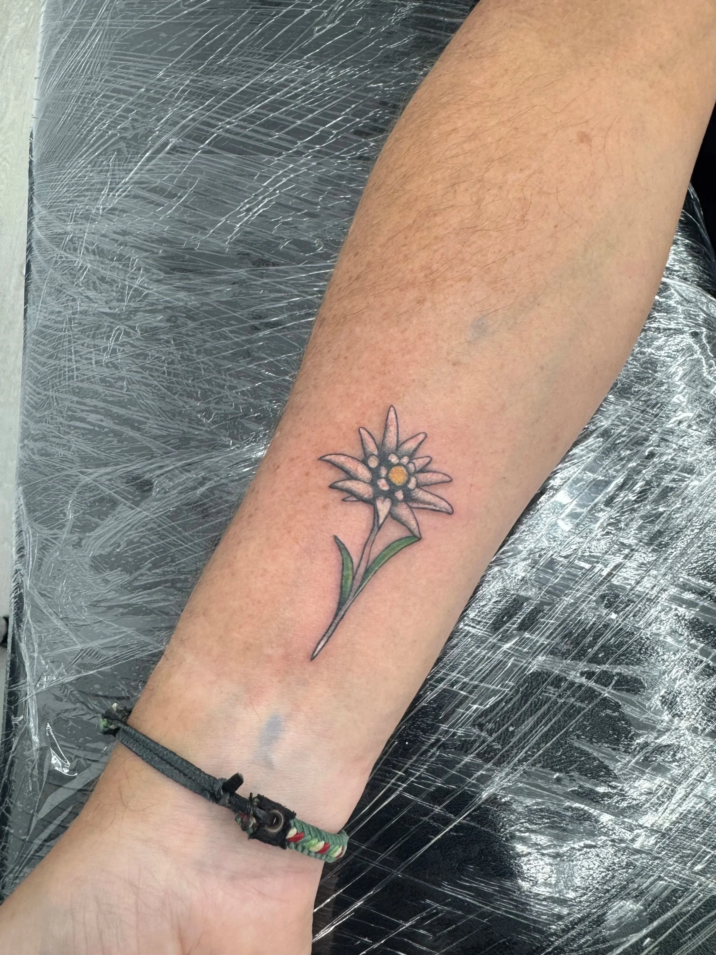 Ein Bein mit einer Tätowierung einer Blume auf der Wade, auf einer schwarzen Oberfläche, umgeben von Kunstleder- oder Plastikfolie.