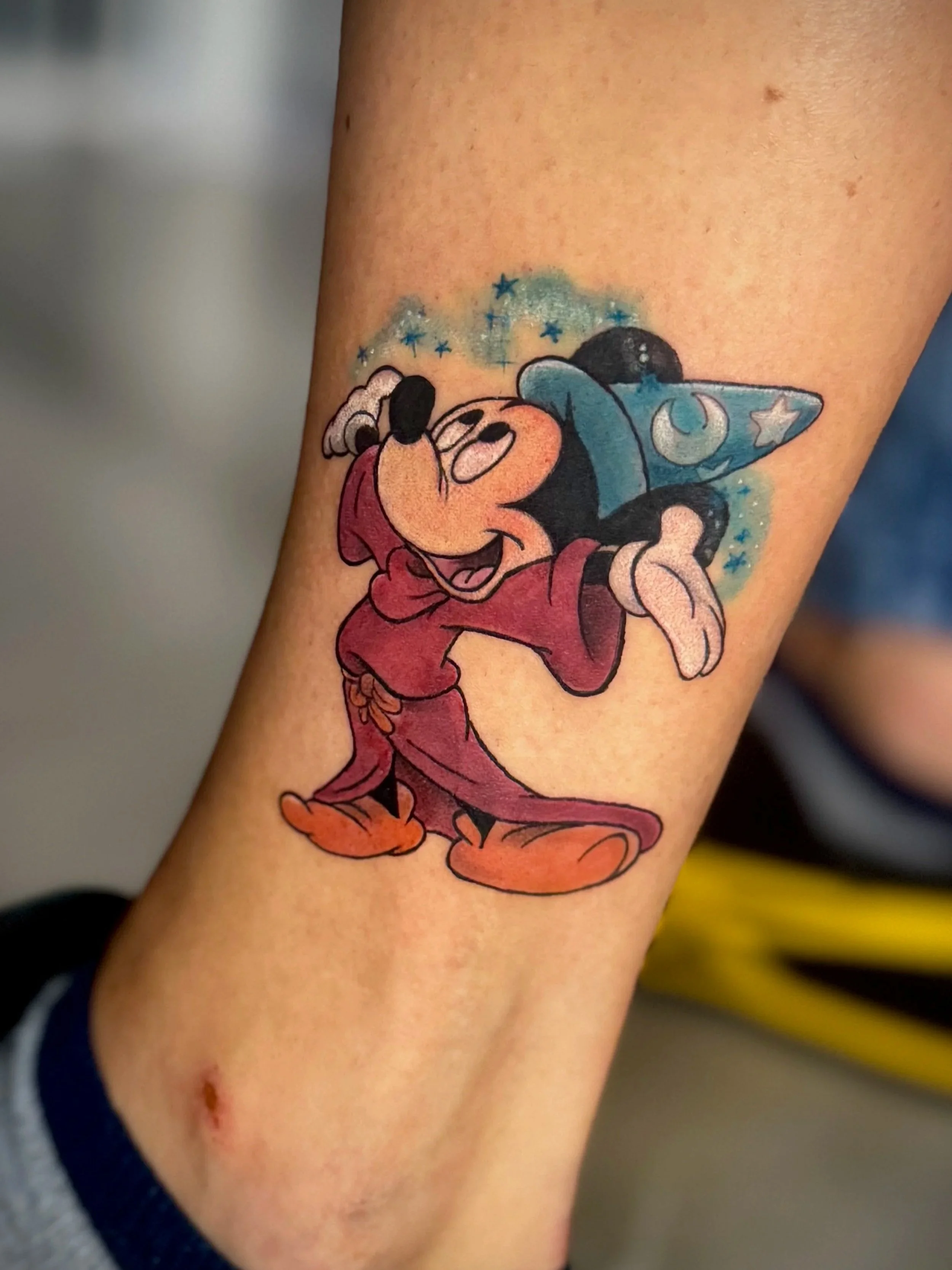 Tattoo von Mickey Mouse, der einen Zauberer-Hut trägt, auf dem Unterarm, mit blauen Sternen und magischer Atmosphäre.