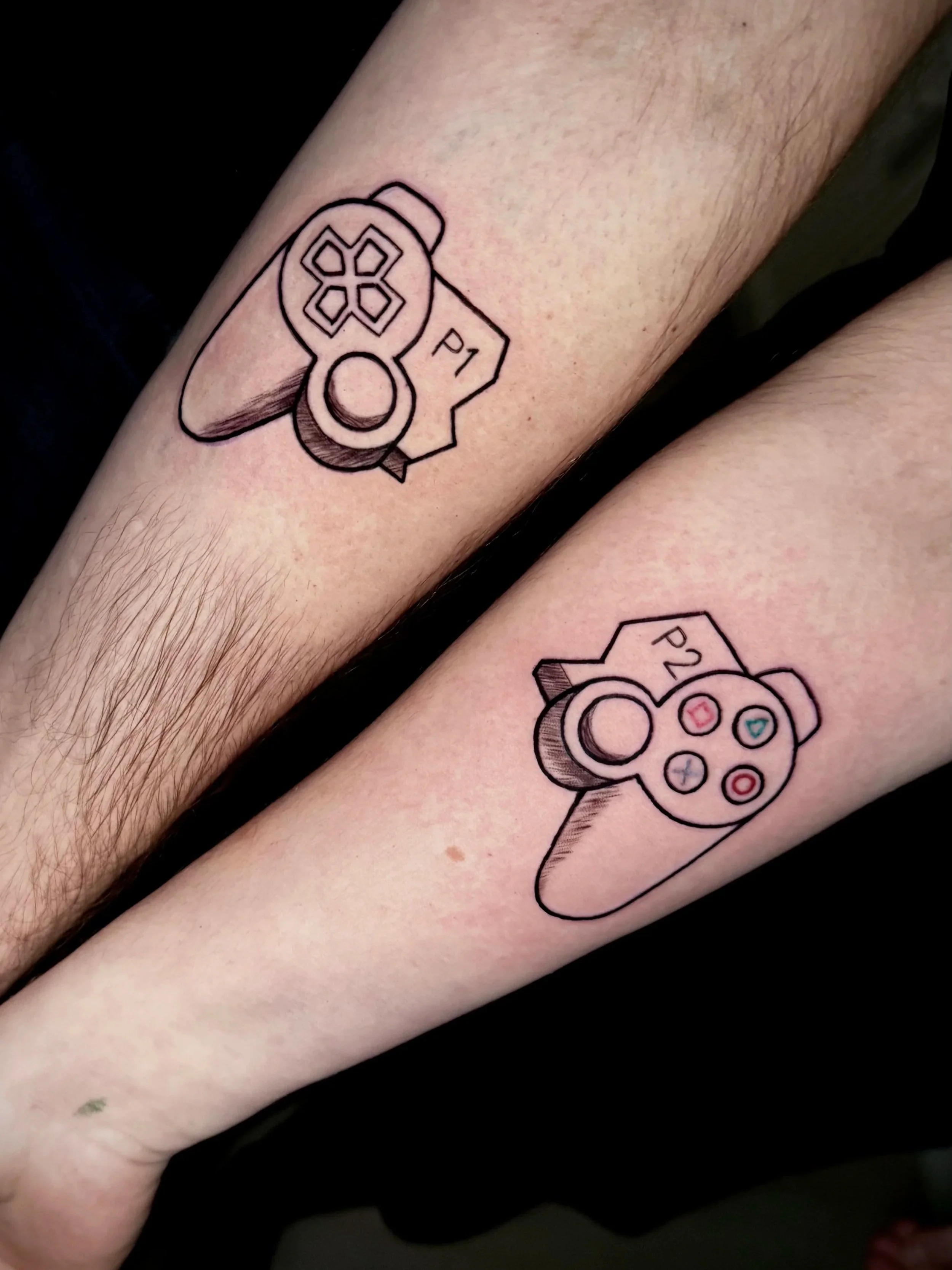 Tattoo von einem PlayStation-Controller auf einem Unterarm, schwarz umrandet, mit farbigen Buttons.