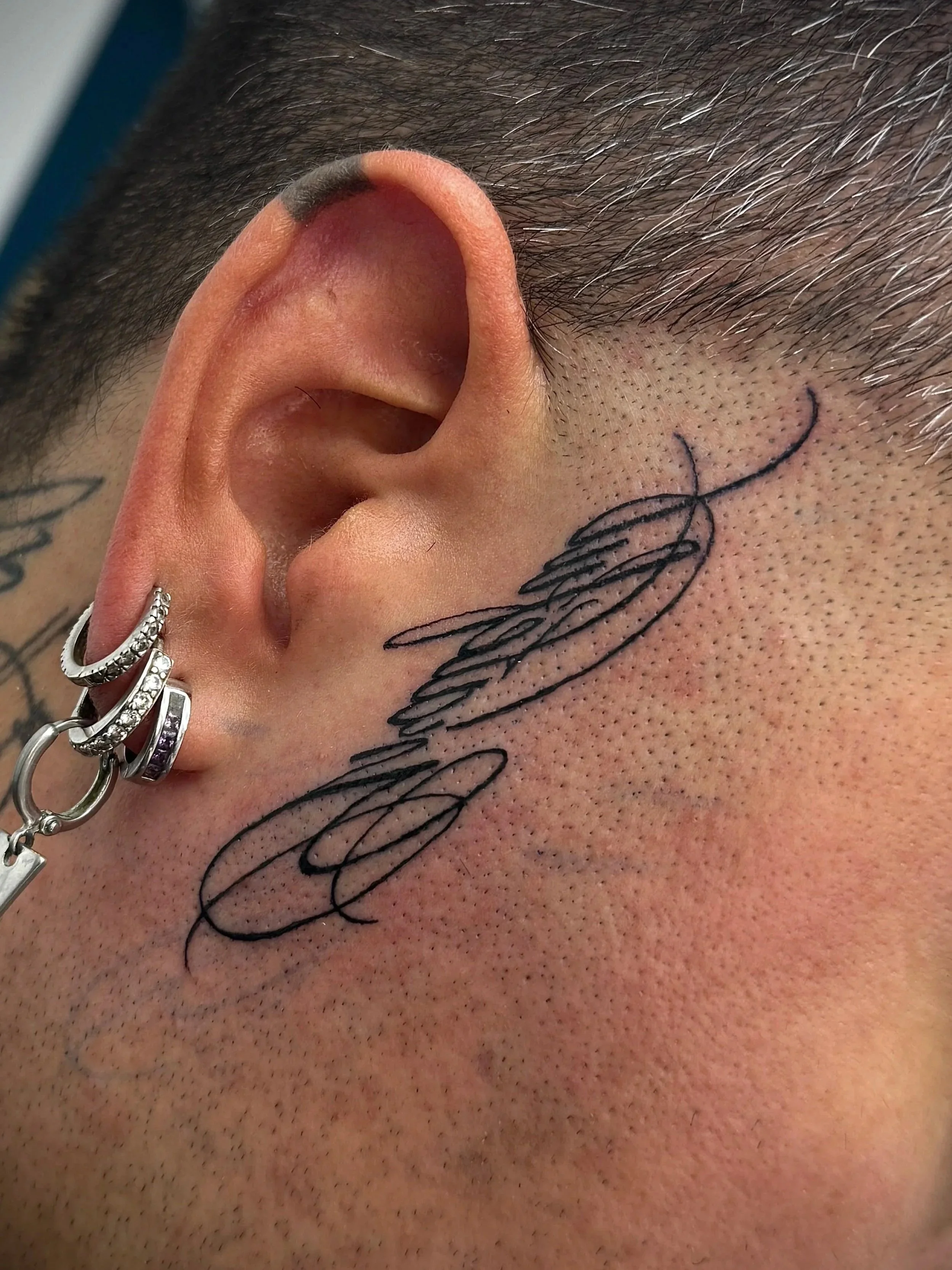 Nahaufnahme eines menschlichen Ohrs mit mehreren Piercings und einem Tattoo auf der Kopfhaut.