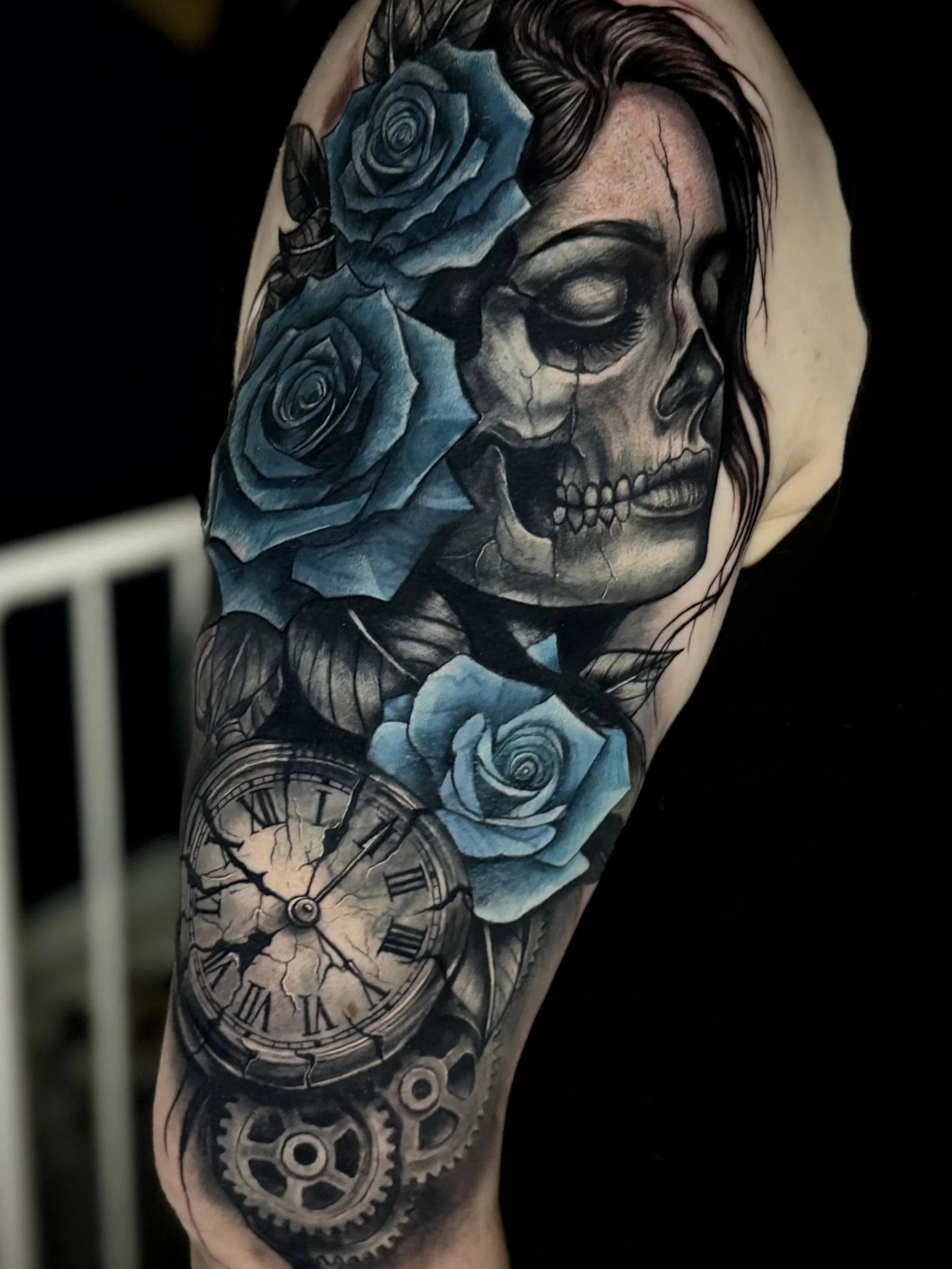 Tattoo eines Schädels mit offenen Kiefern, umgeben von blauen Rosen, mit Uhr und Zahnrädern auf einem Arm.