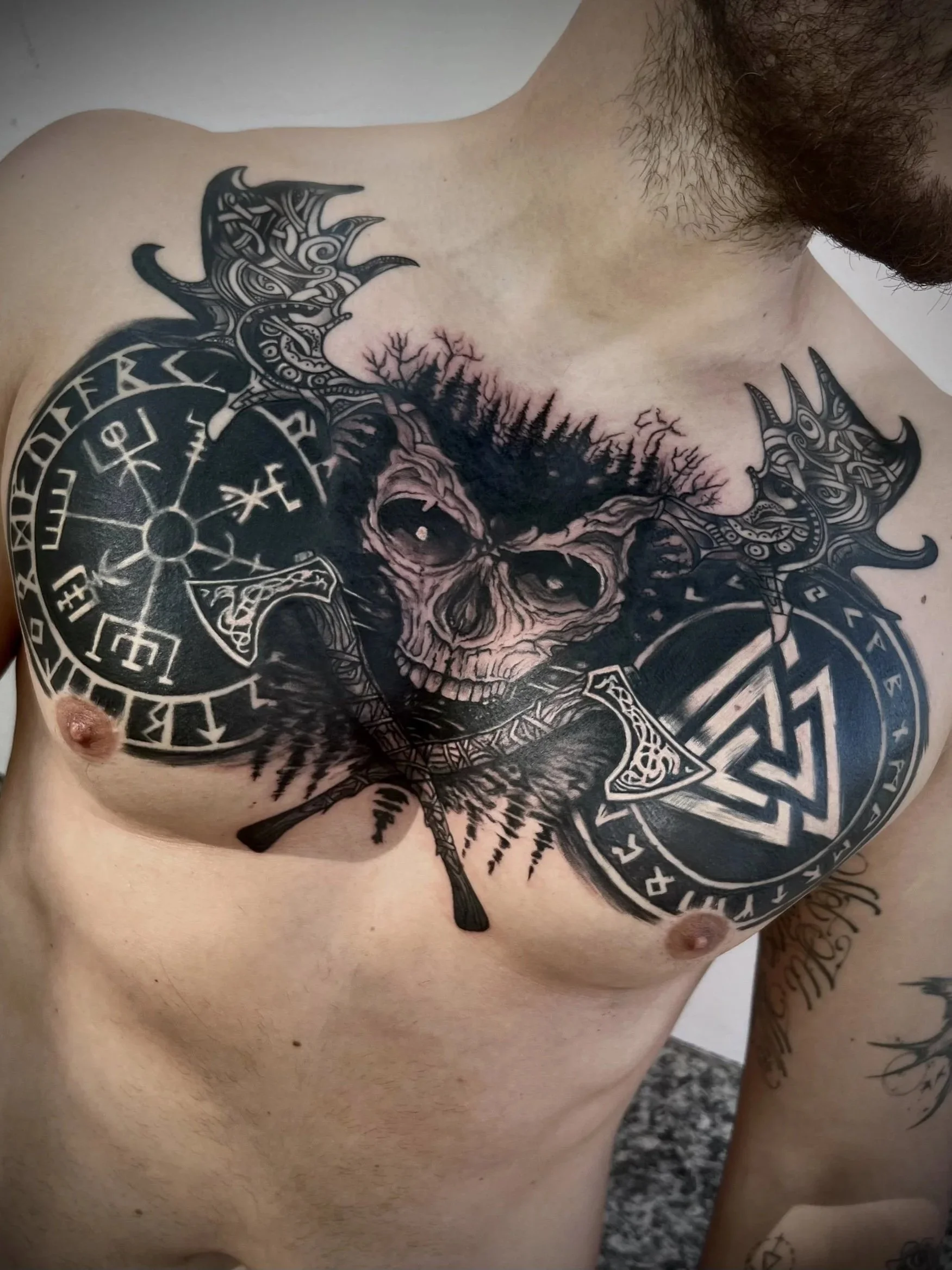 Tattoo eines Totenkopfes mit Raben und Runen auf der Brust eines Mannes.
