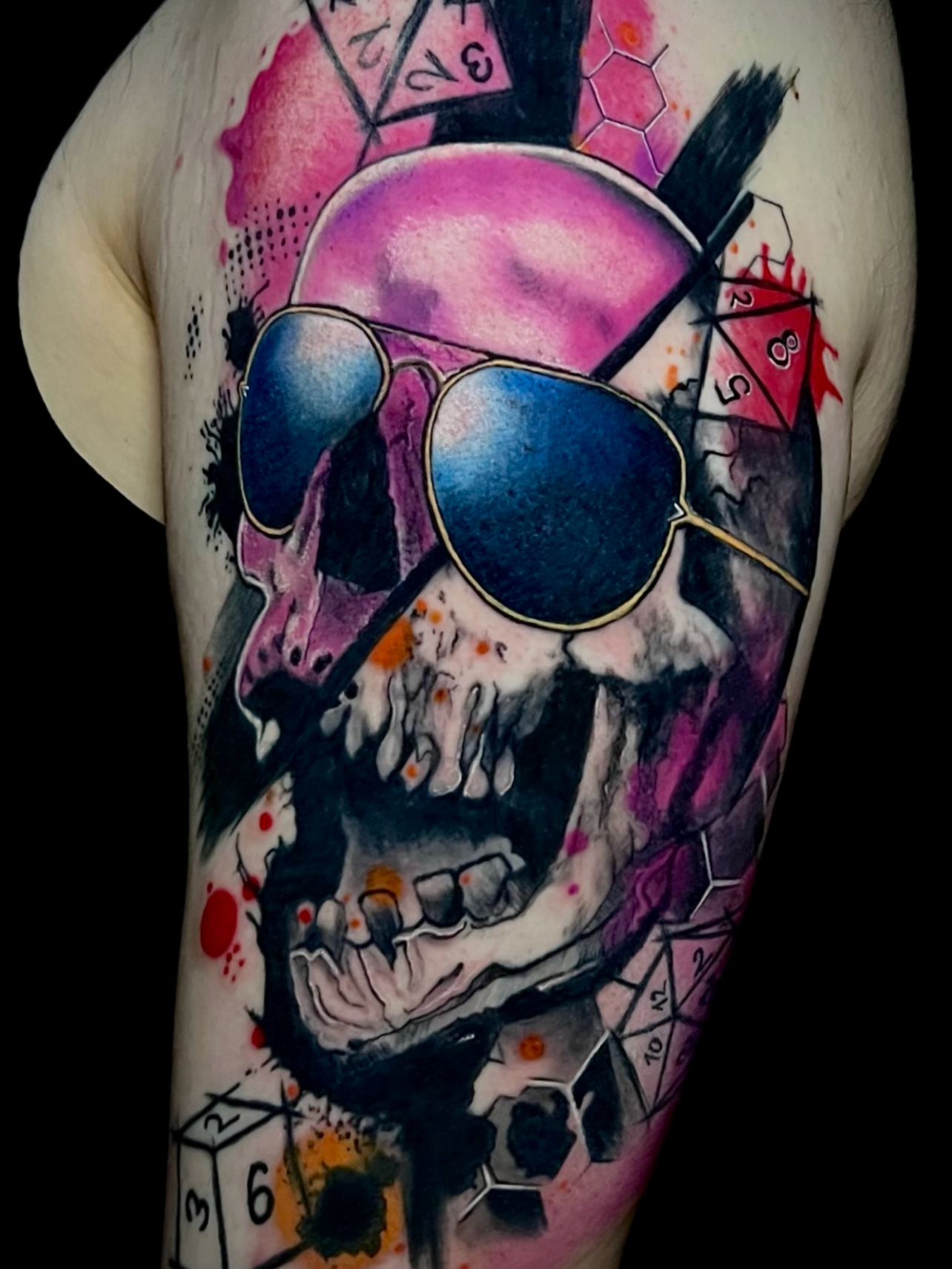Ein bärtiger Mann mit schwarzem Haar trägt eine bunte Tattoo auf seinem Arm, das ein menschliches Skelett, ein Pinkes Gesicht mit Sonnenbrille, Schwarze Federn, Würfel und andere geometrische Formen zeigt.