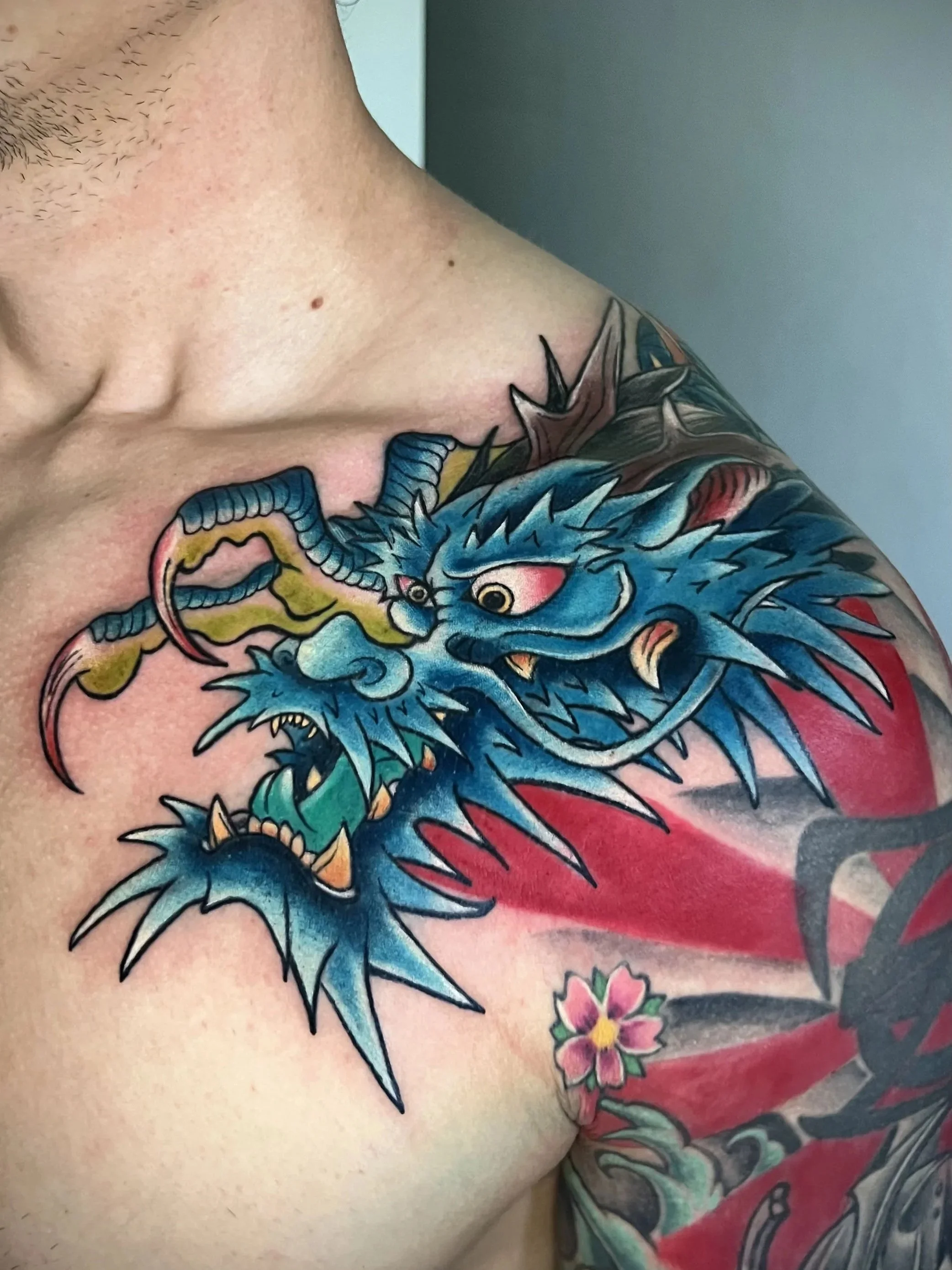 Nahaufnahme eines Tattoos eines blauen und goldenen chinesischen Drachen mit Pinkfarbenen Akzenten auf der Schulter eines Menschen, umgeben von weiteren farbenfrohen Tattoos und einem Blumenmotiv.