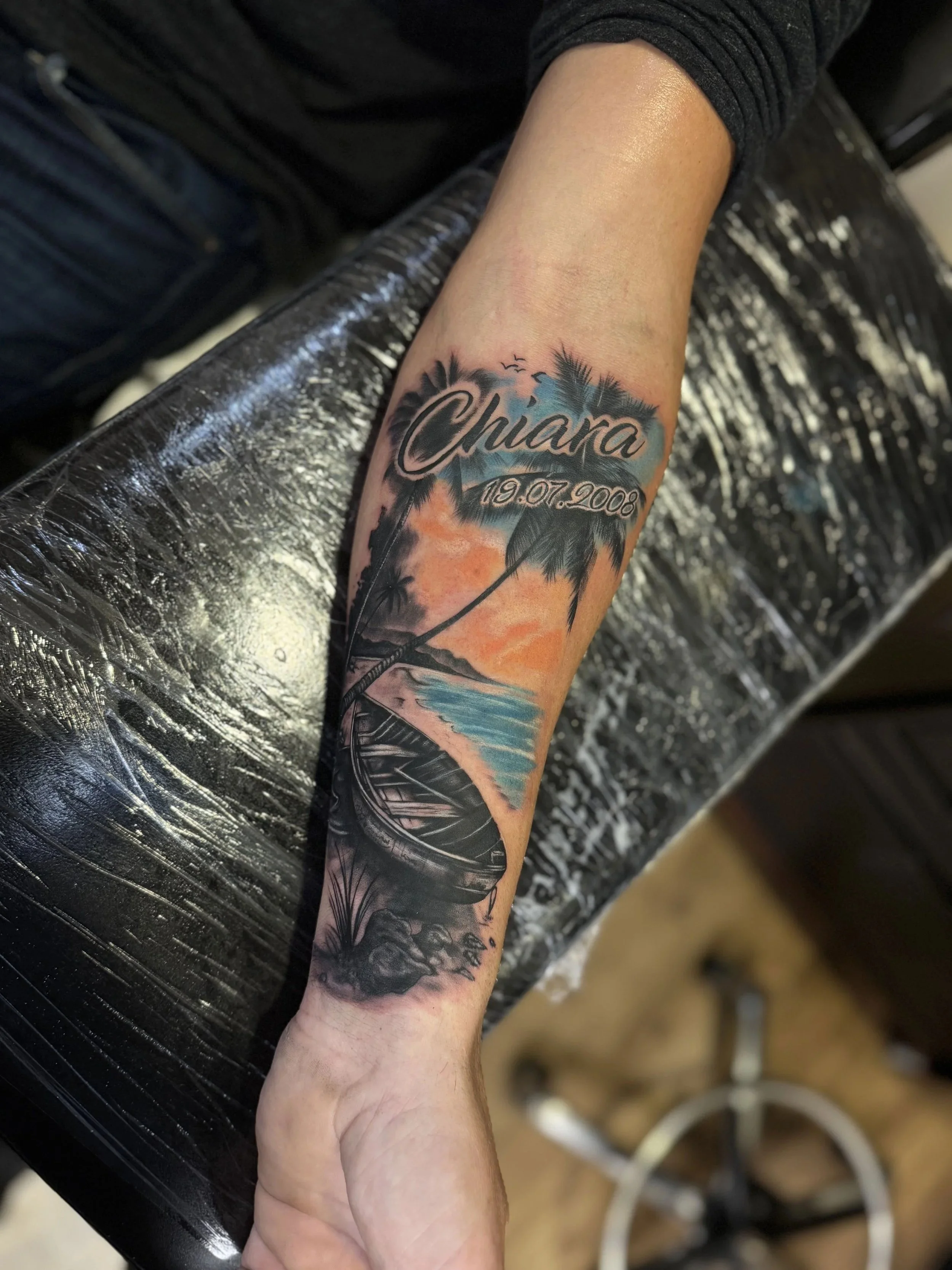 Ein Arm mit einem farbenfrohen Tattoo, das Palmen, einen Sonnenuntergang und einen Himmel zeigt. Der Name »Chiara« und das Datum »19.07.2008« sind eingebettet.