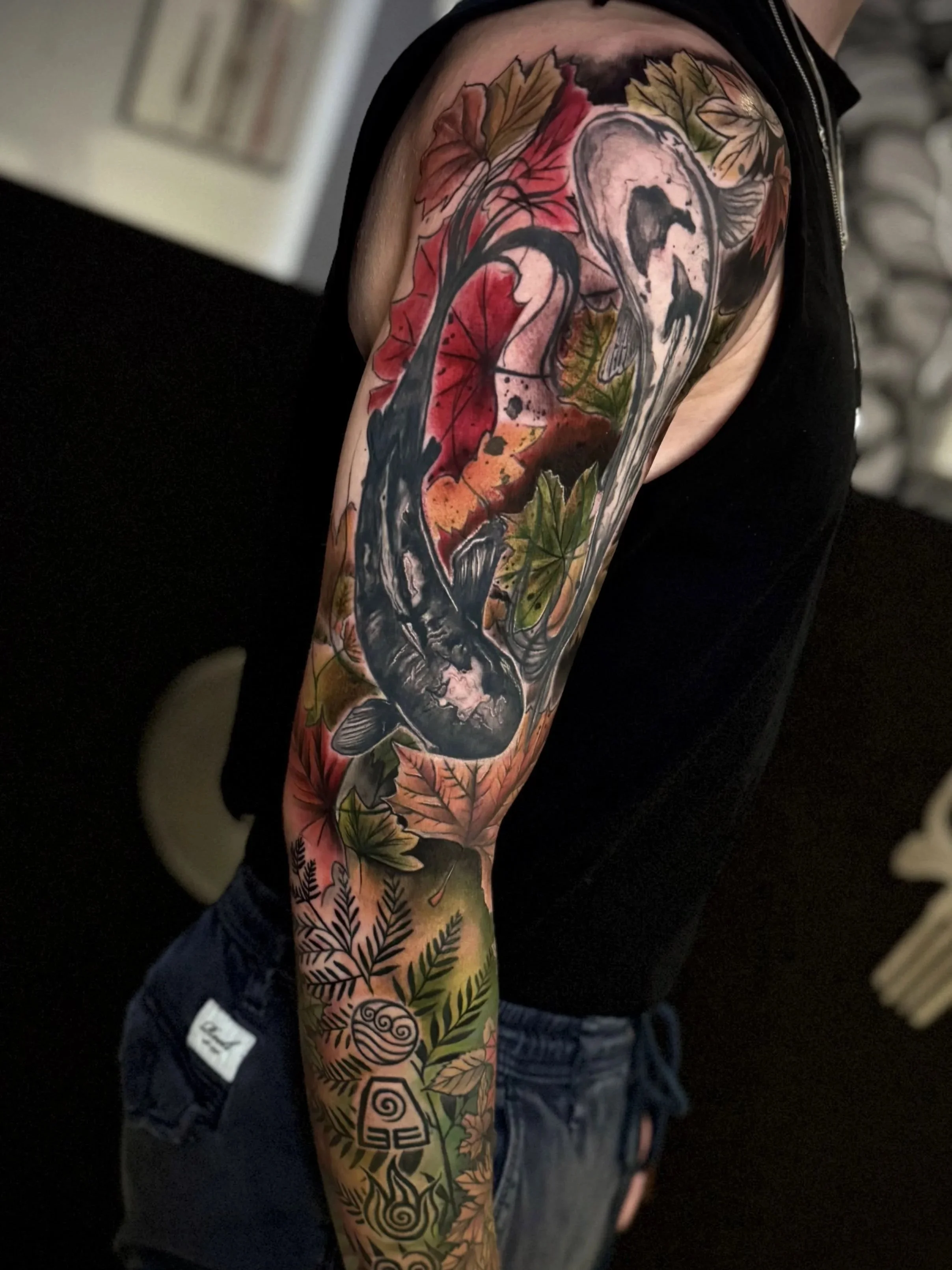 Nahaufnahme eines Tattoos auf dem Arm einer Person, das verschiedene bunte Blätter und ein schwarzer Schwan zeigt.