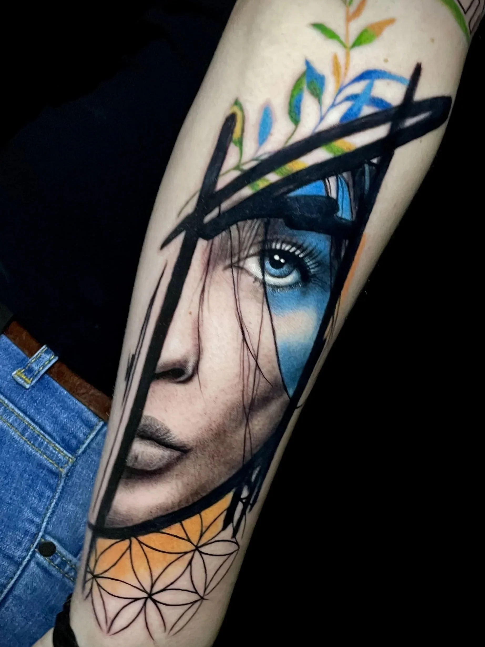Tätowierung eines menschlichen Gesichts mit blauen Augen, schwarzen Haaren und detailliertem Gesichtsausdruck auf einem Arm, ergänzt durch geometrisches und Blumendesign.