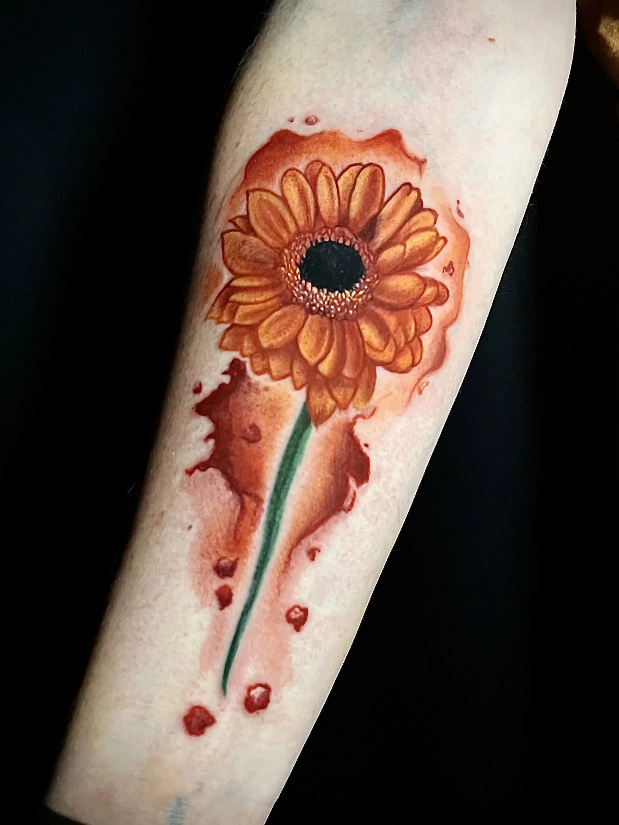 Tattoo eines orangefarbenen Blumenmusters mit schwarzem zentralem Kreis auf einem menschlichen Arm.