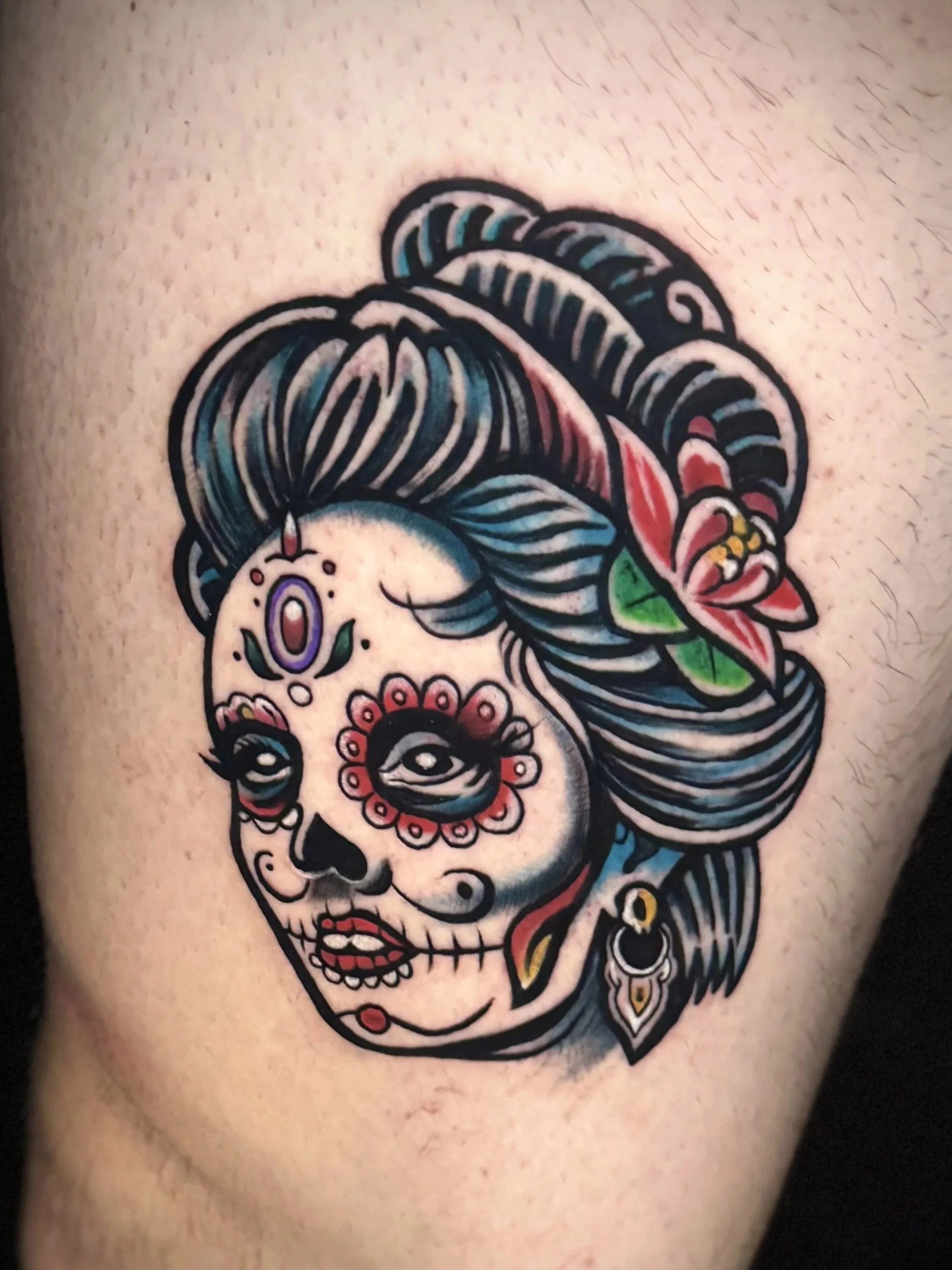 Eine bunte Tattoo-Darstellung eines Totenkopfes mit geometrischen und floralen Mustern, verziert mit einer Figur, die an eine historische Tänzerin erinnert.