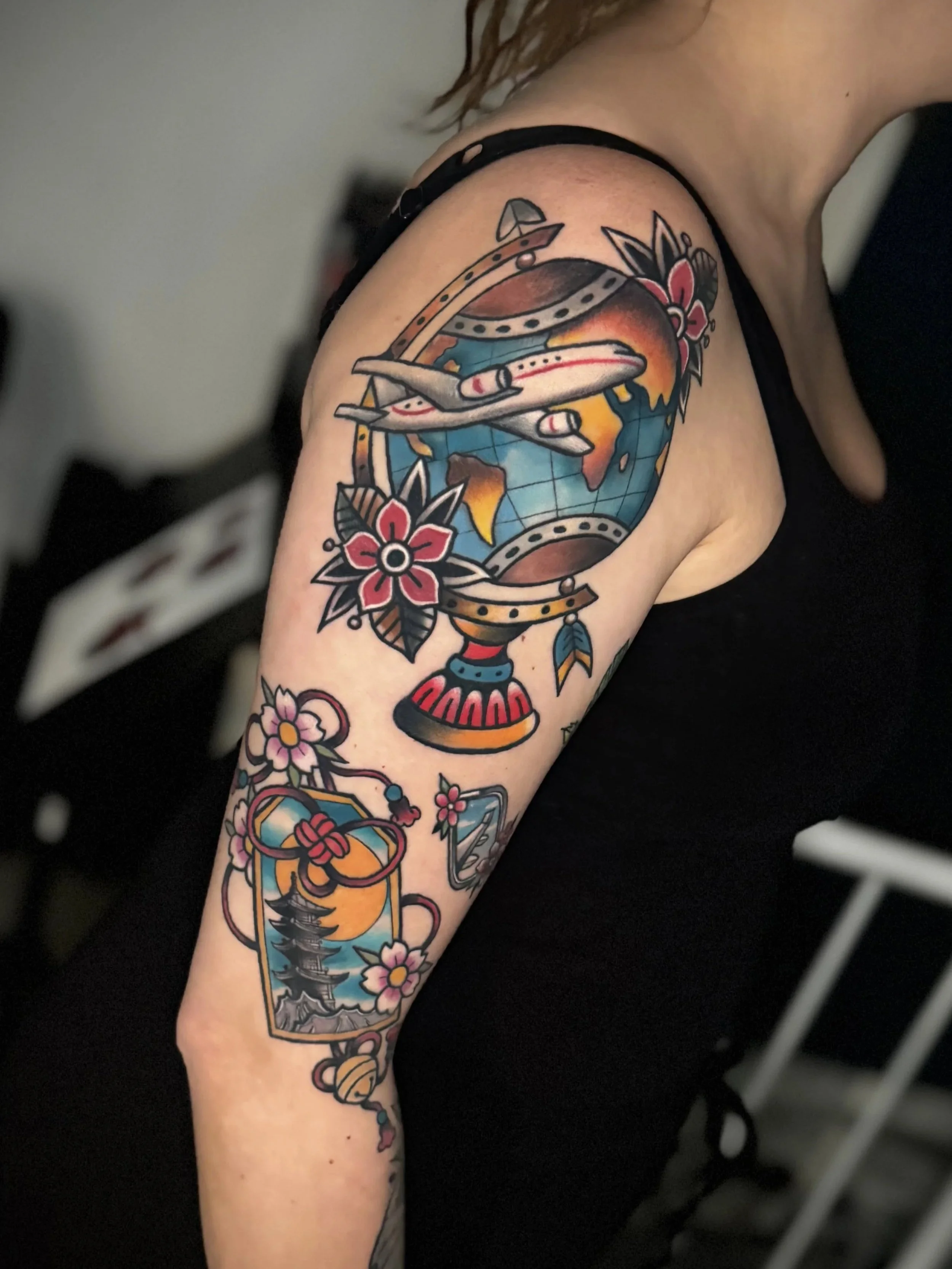 Tattoo auf dem Oberarm einer Frau, das eine Weltkarte, eine Flugzeug-Silhouette, Blumen und traditionelle Tattoo-Designs zeigt.