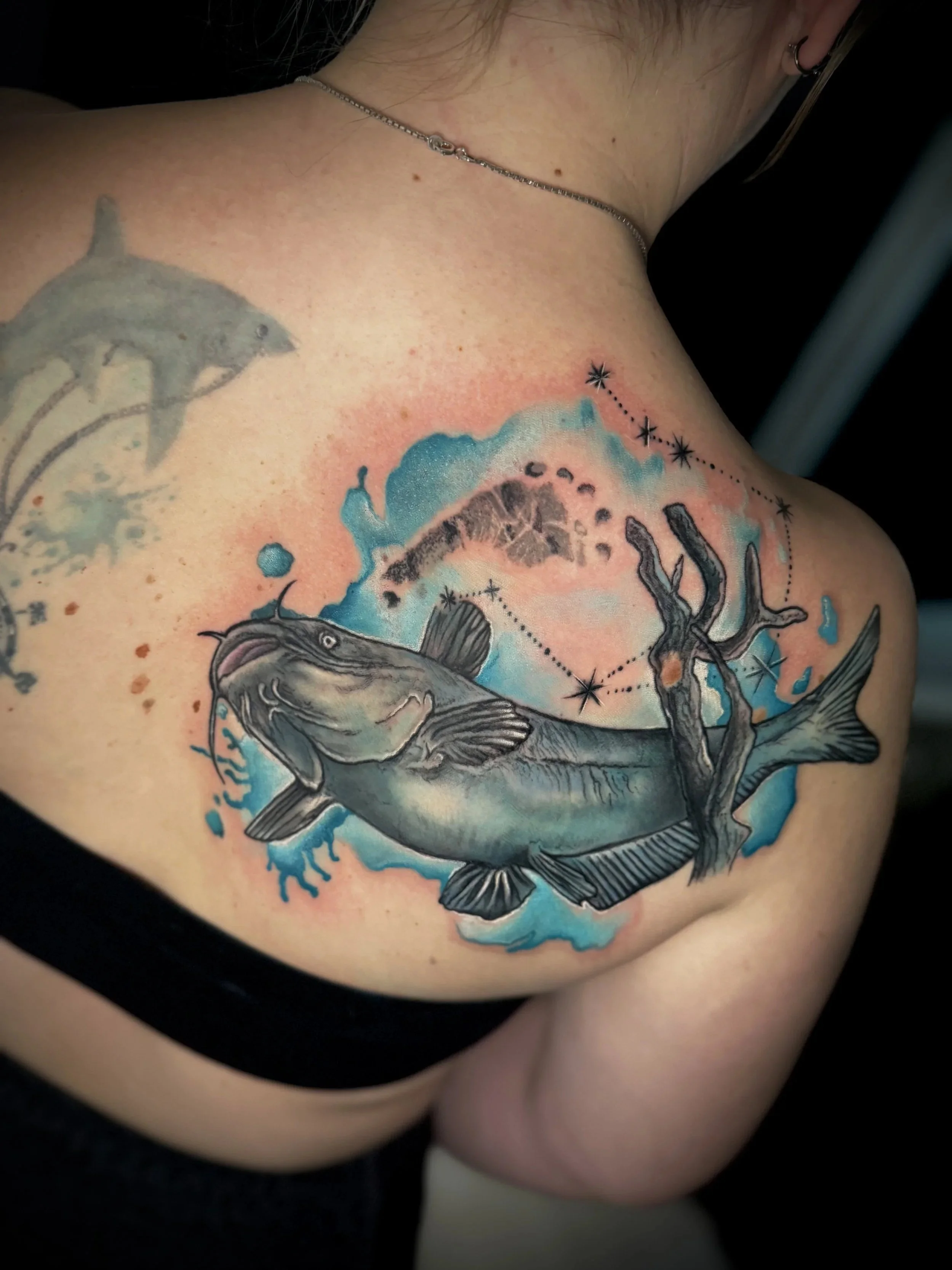 Tattoo eines Fisches auf der Schulter einer Person, mit blauer Wasserfarbe und Sternenkonstellation in der Nähe.