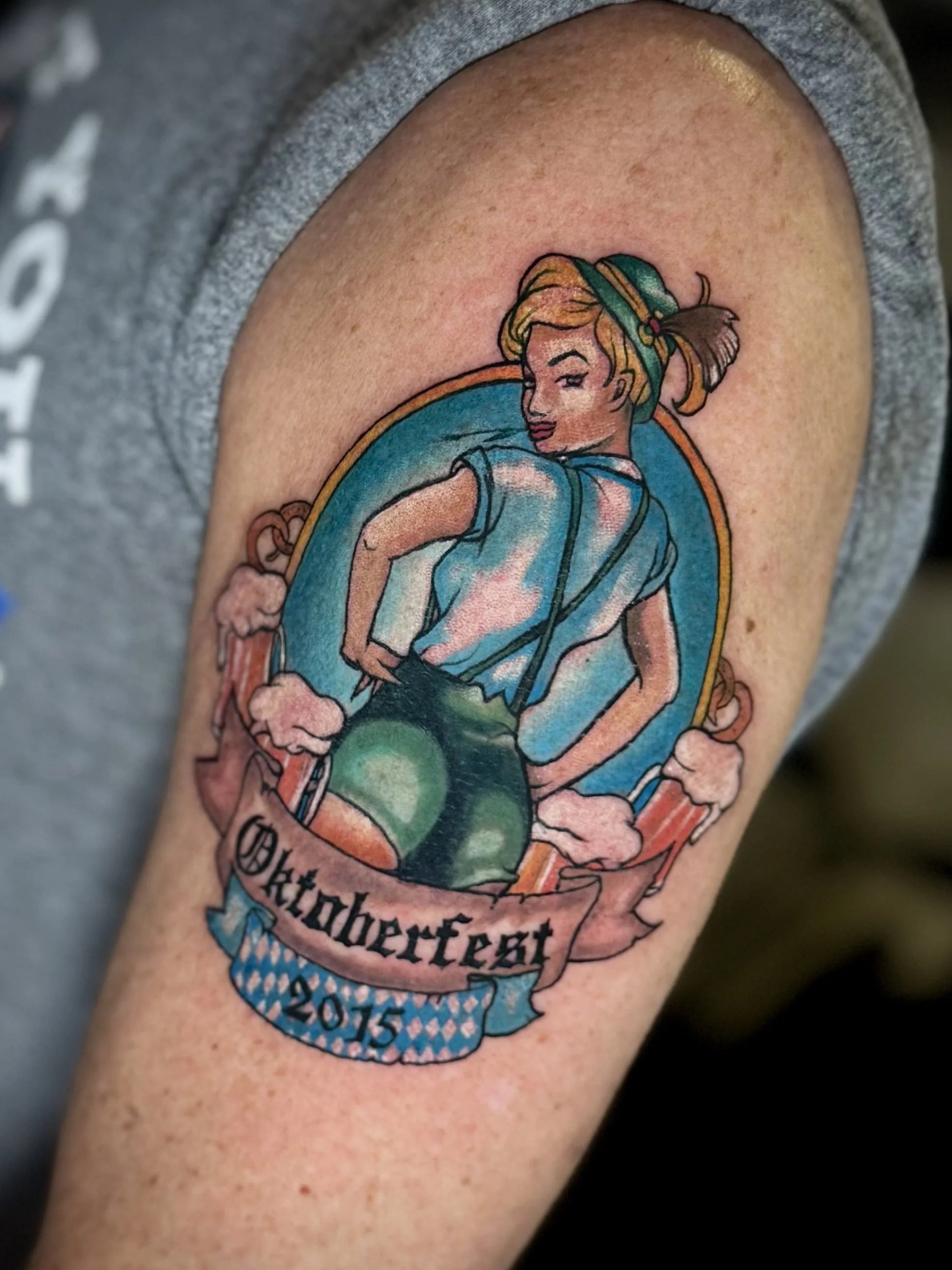 Tattoo eines Mädchens in dirndlähnlicher Kleidung mit Hut, das in einem Kreis sitzt, mit deutschem Text und der Jahreszahl 2015.