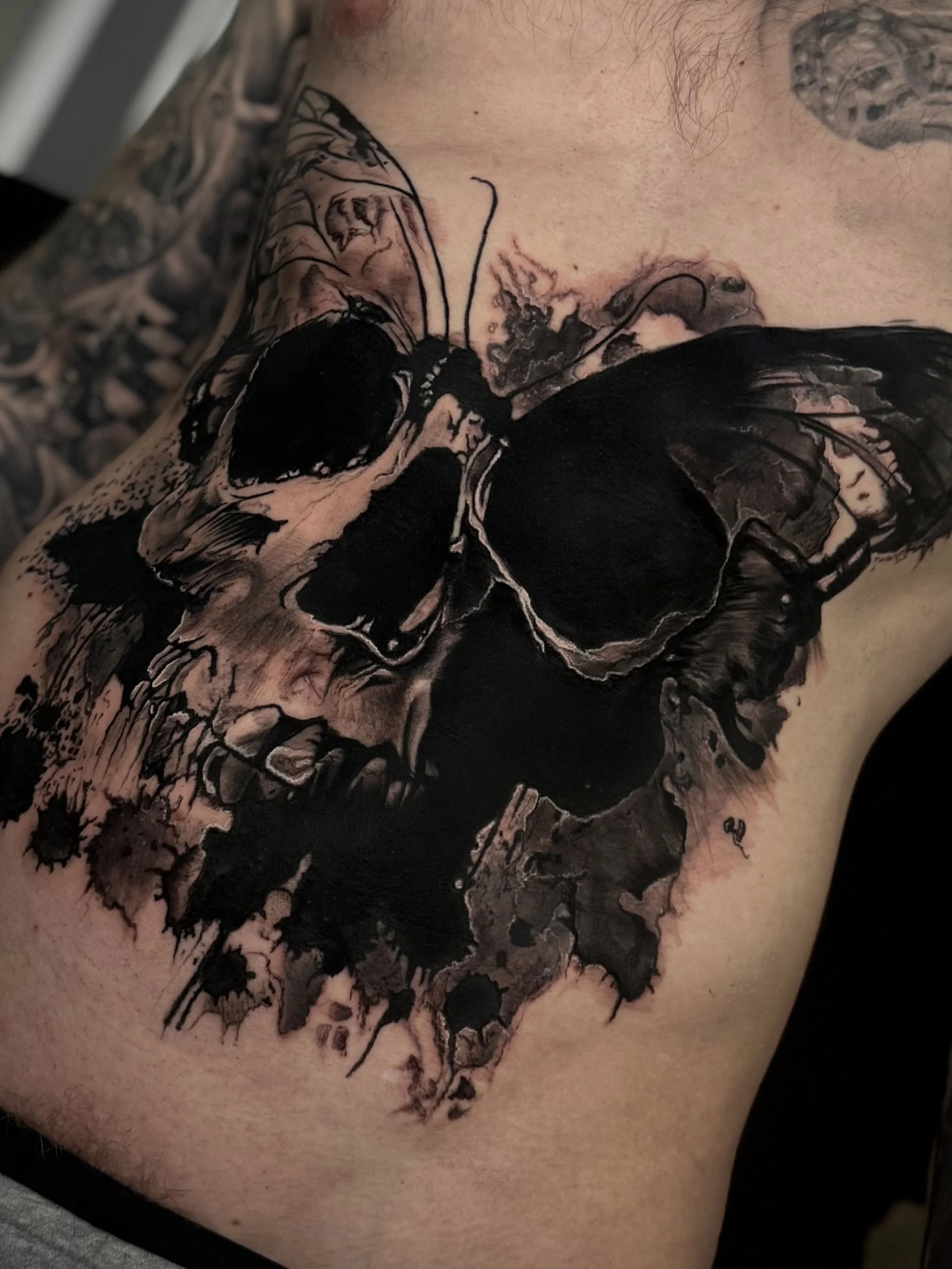 Tattoo eines schwarzen Schmetterlings auf der Schulter mit schwarzen, aquarellartigen Akzenten.