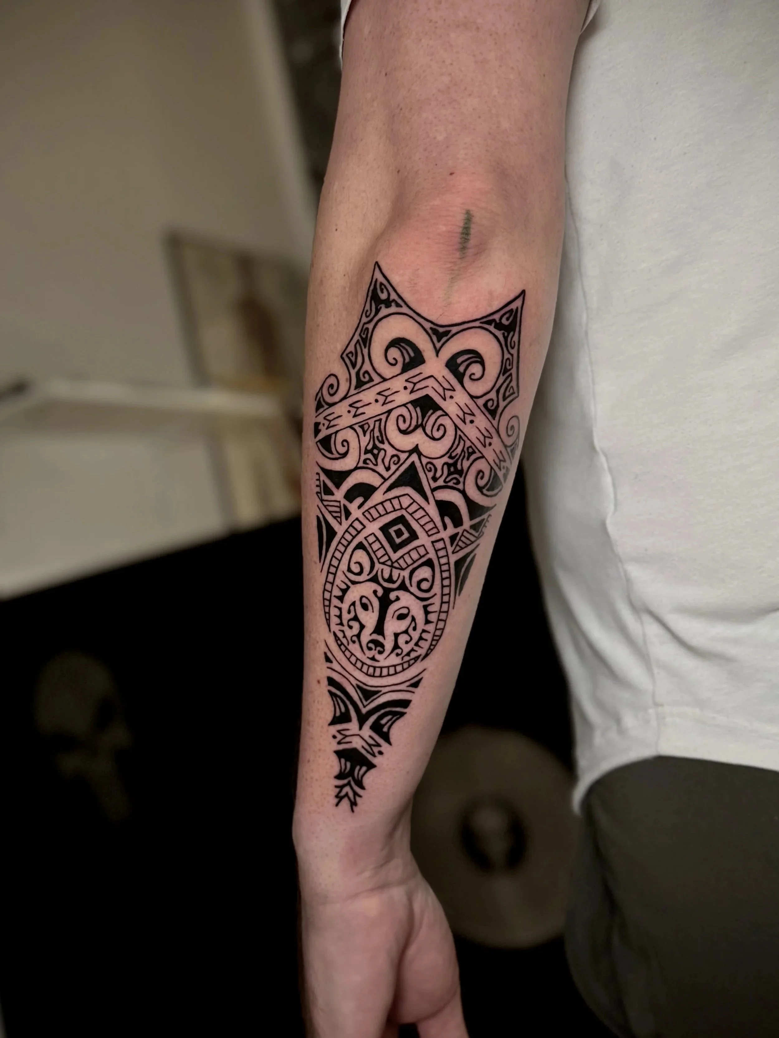 Der Bild zeigt einen menschlichen Arm mit einem traditionellen schwarzen Tribal-Tattoo. Das Tattoo ist detailliert mit Mustern und Linien, die sich entlang des Unterarms erstrecken.