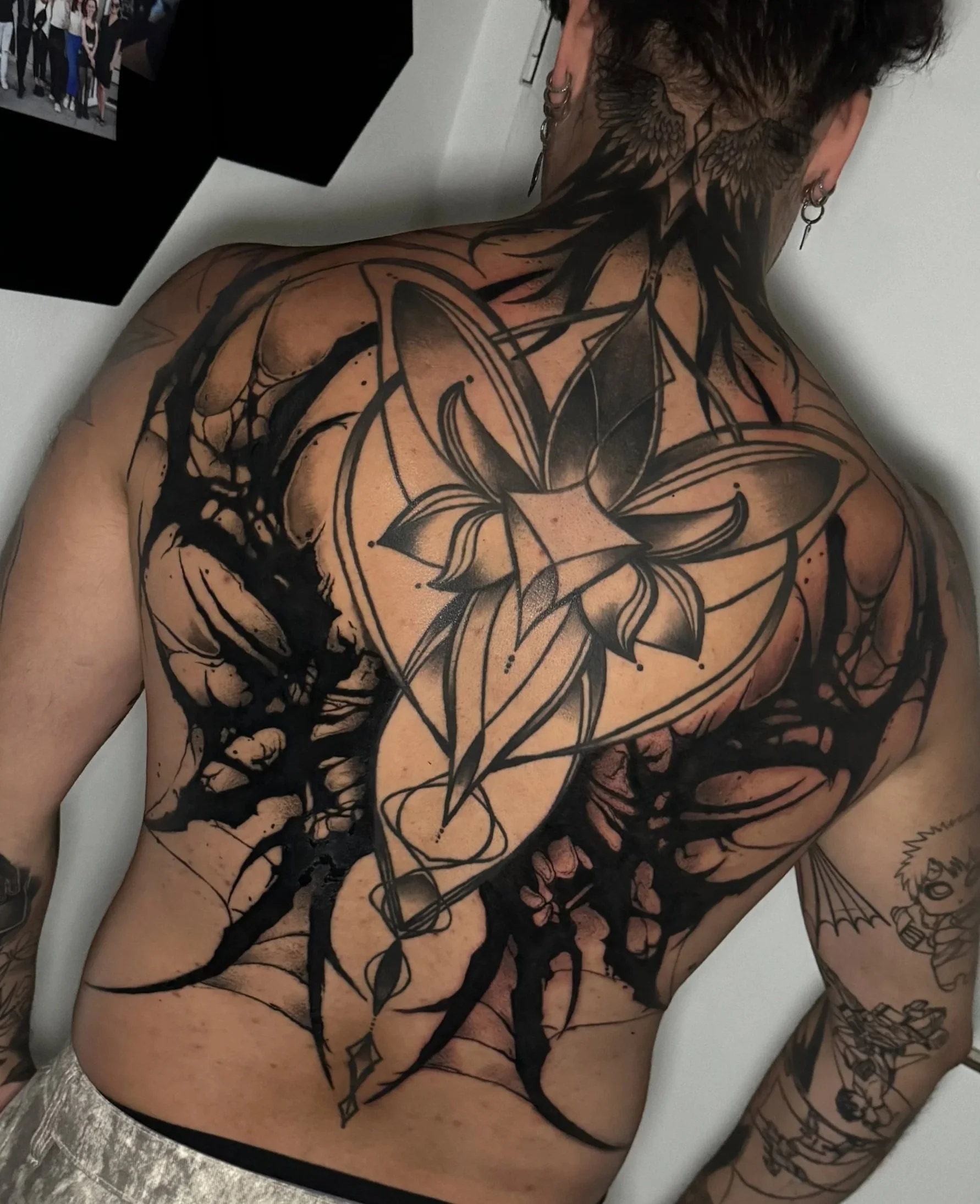 Nackte Person mit Tattoos auf Rücken und Arm, sitzt auf einem Stuhl, Blick nach unten, im Hintergrund ein Bildschirm mit einem Gruppenfoto, dezenten Schmuck an den Ohren, schwarzes Haar.