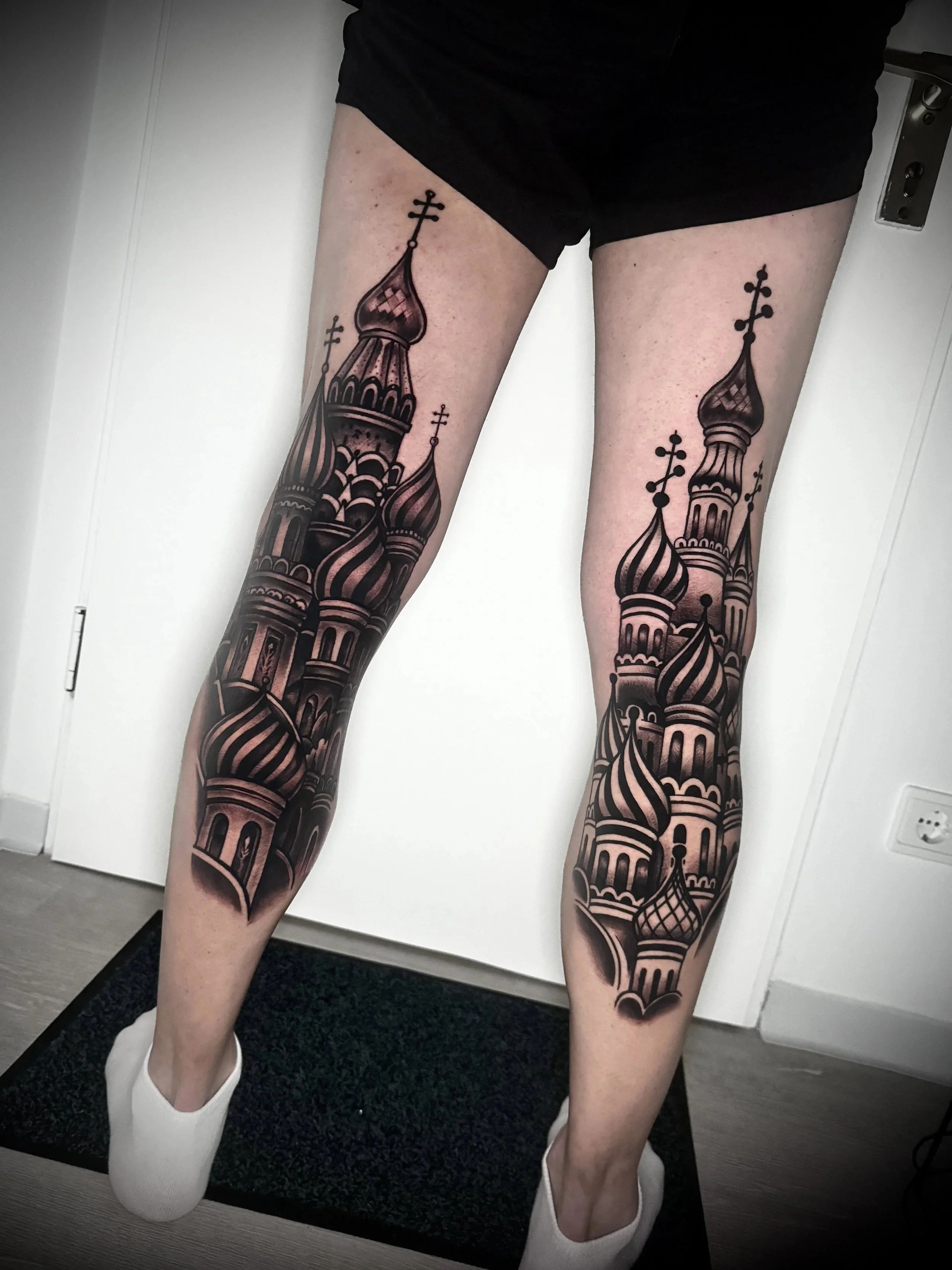 Tattoo von russischen Kathedralen auf den Beinen einer Person in schwarzen Shorts, die auf einem schwarzen Teppich sitzt, mit weißen Socken und weißen Schuhen, vor einer weißen Tür.