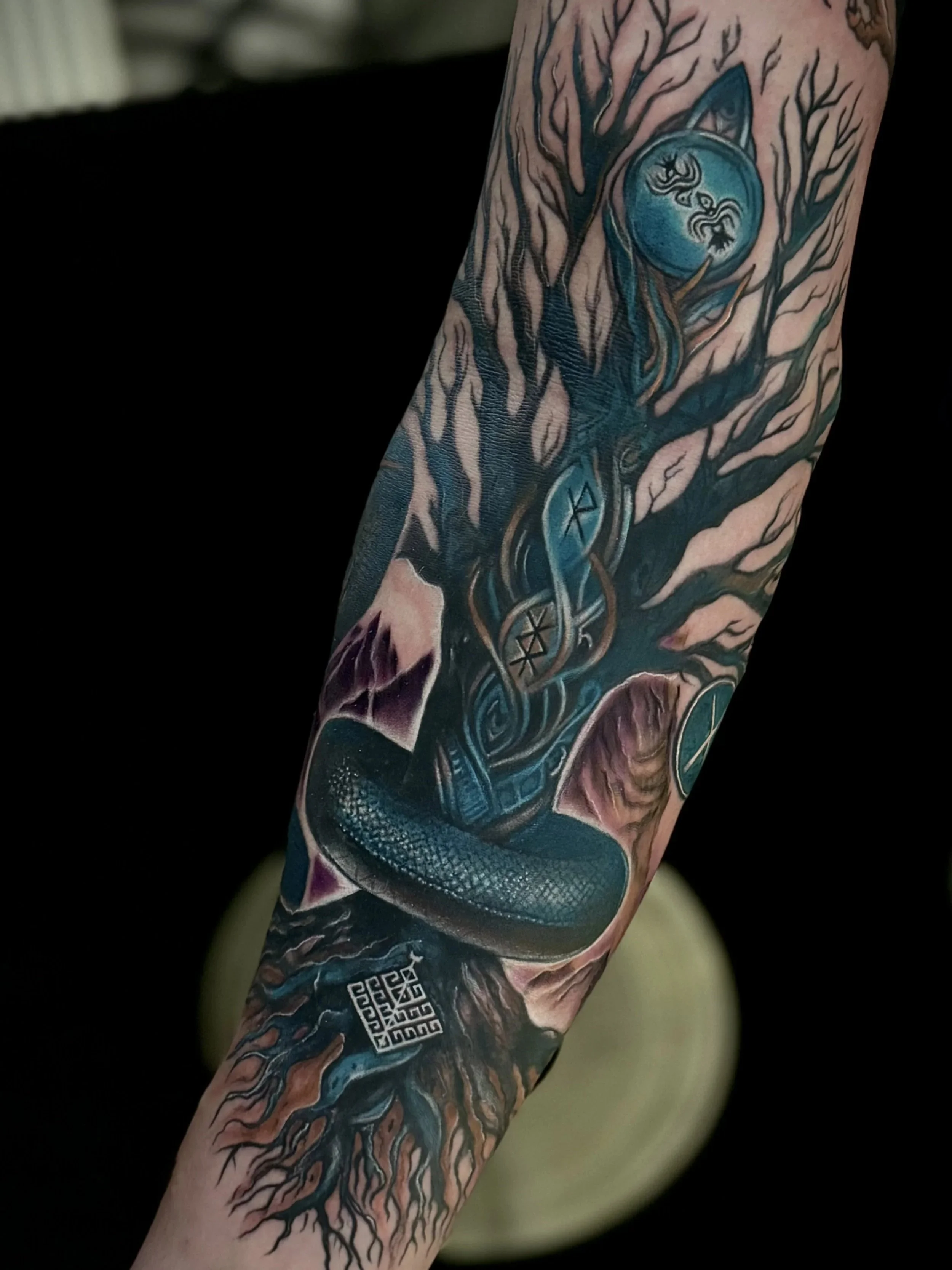 Detailliertes Tattoo eines Baumes mit knorrigen Ästen, einer blauen Mondscheibe mit Drachen und Ringen, sowie QR-Codes und chinesischen Symbolen auf dem Unterarm.