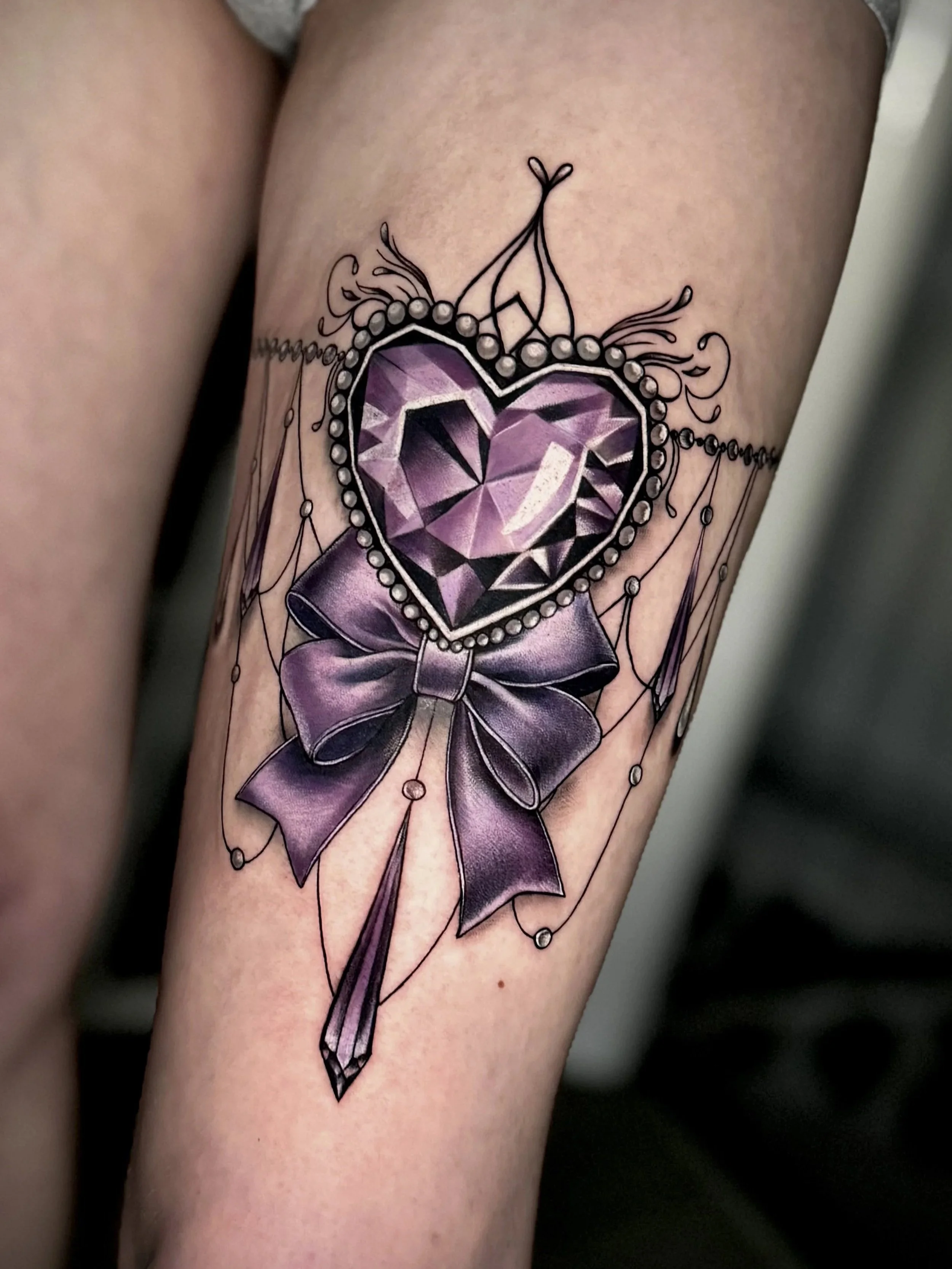 Taillentattoo eines herzförmigen, violett schimmernden Edelsteins mit violettem Schleifenband und Perlen, umgeben von filigranen schwarzen Linien.