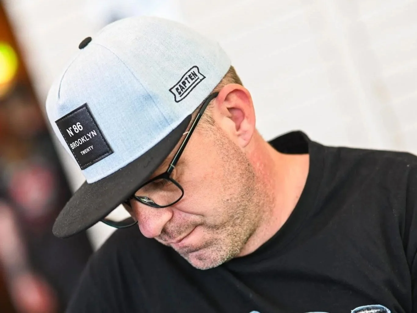 Ein Mann trägt eine Baseballkappe mit Aufnähern, liest oder schreibt, trägt Brille, trägt einen schwarzen Pullover.
