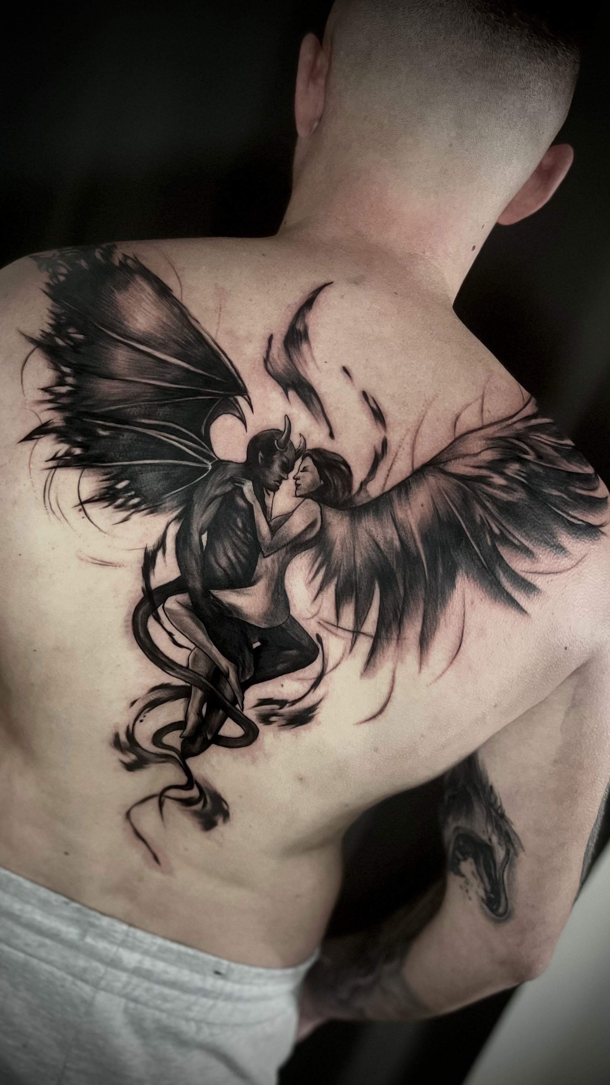 Dunkle Tattoo auf Schulter und Oberarm eines Mannes, zeigt eine Teufelin mit Flügeln und einem Gegenüber, der sich berühren.