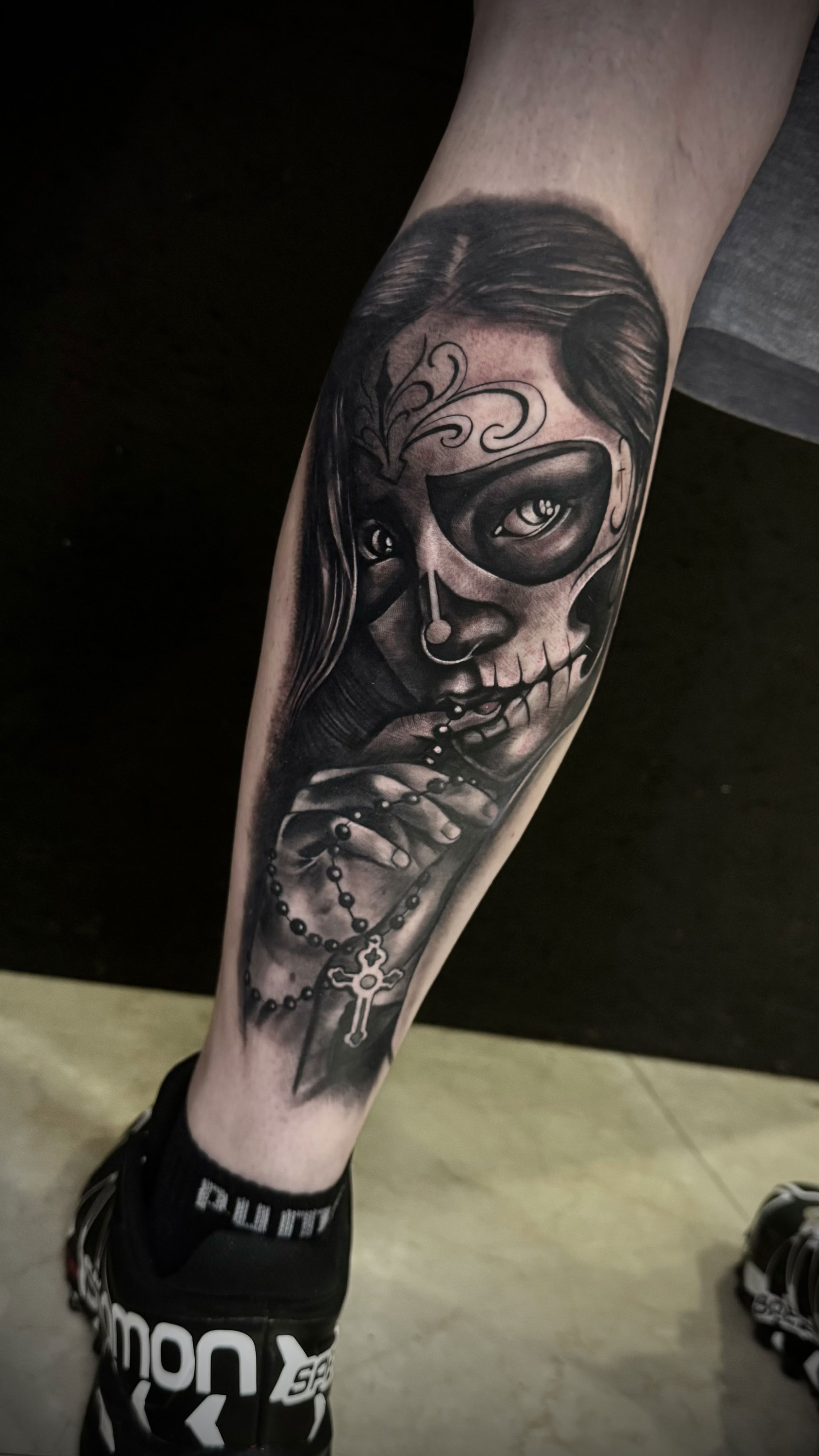 Schwarz-weiße Tattoo eines Mädchens mit Geistergesicht, mit Halskette und Kreuz. Das Tattoo ist auf dem Unterschenkel sichtbar.