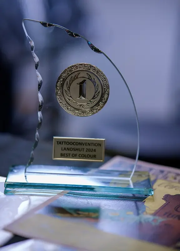 Trophäe mit einer Medaille, die auf einer transparenten Basis sitzt, darunter eine Beschriftung mit 'TattooConvention Landshut 2024 Best of Colour', auf einem Tisch mit bunten Papieren.