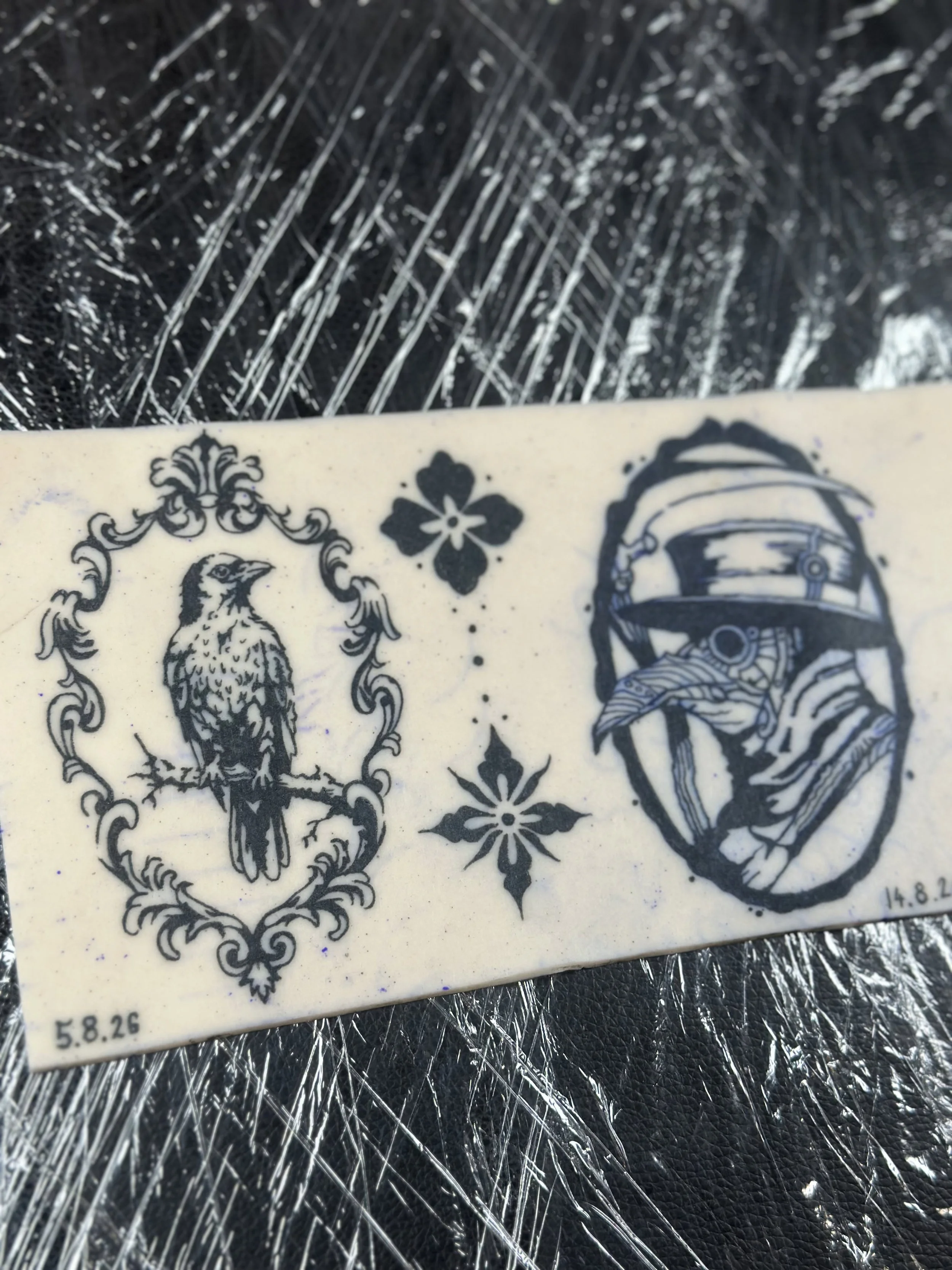 Ein Kachel mit schwarzen und weißen grafischen Designs, darunter ein Vogel, eine Blume und ein Helm. Das Bild liegt auf einer auffällig strukturierten schwarzen Oberfläche.