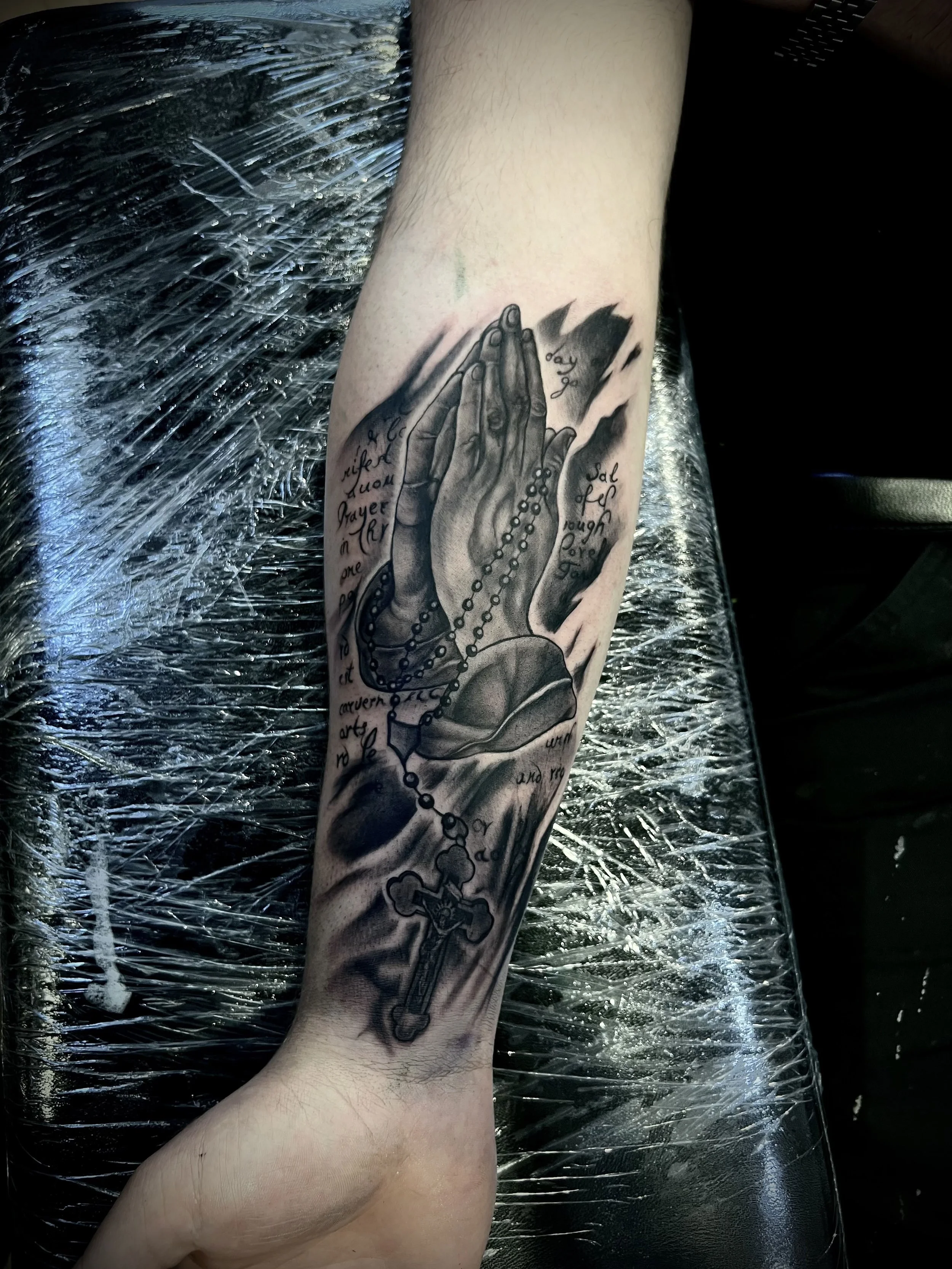 Tattoo auf dem Unterarm einer Person, das betende Hände mit Gebetskette zeigt, umgeben von französischer und englischer Schrift. Die Person liegt auf einer Schutzfolie, wahrscheinlich beim Tätowieren.