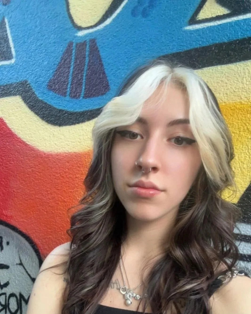 Ein junges Mädchen mit braunen, gewellten Haaren, das vor einer bunten, graffitiartigen Wand steht.