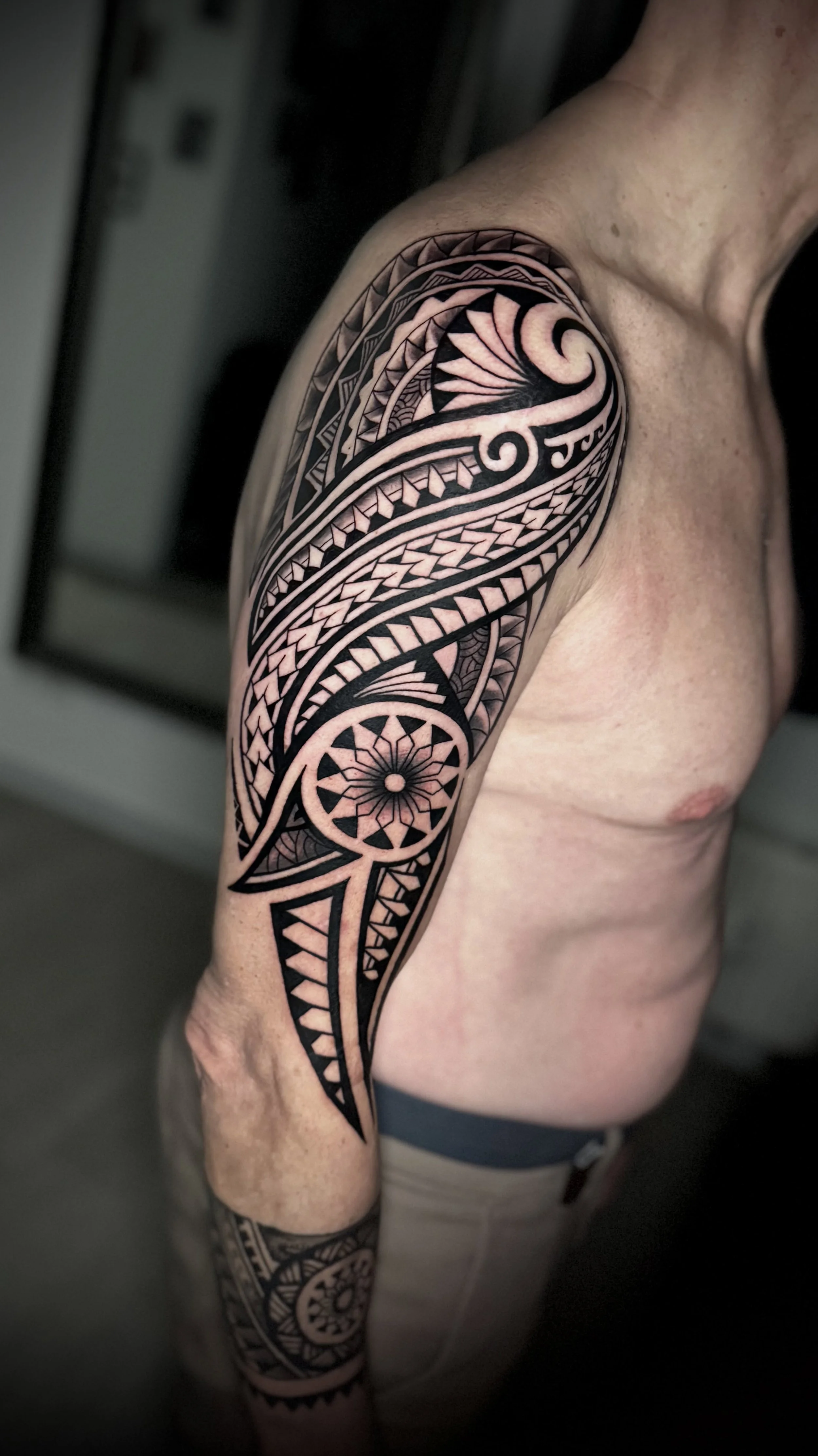 Schwarzes Tribal-Tattoo auf dem Oberarm und Unterarm eines Mannes, mit geometrischen und spiralförmigen Mustern.