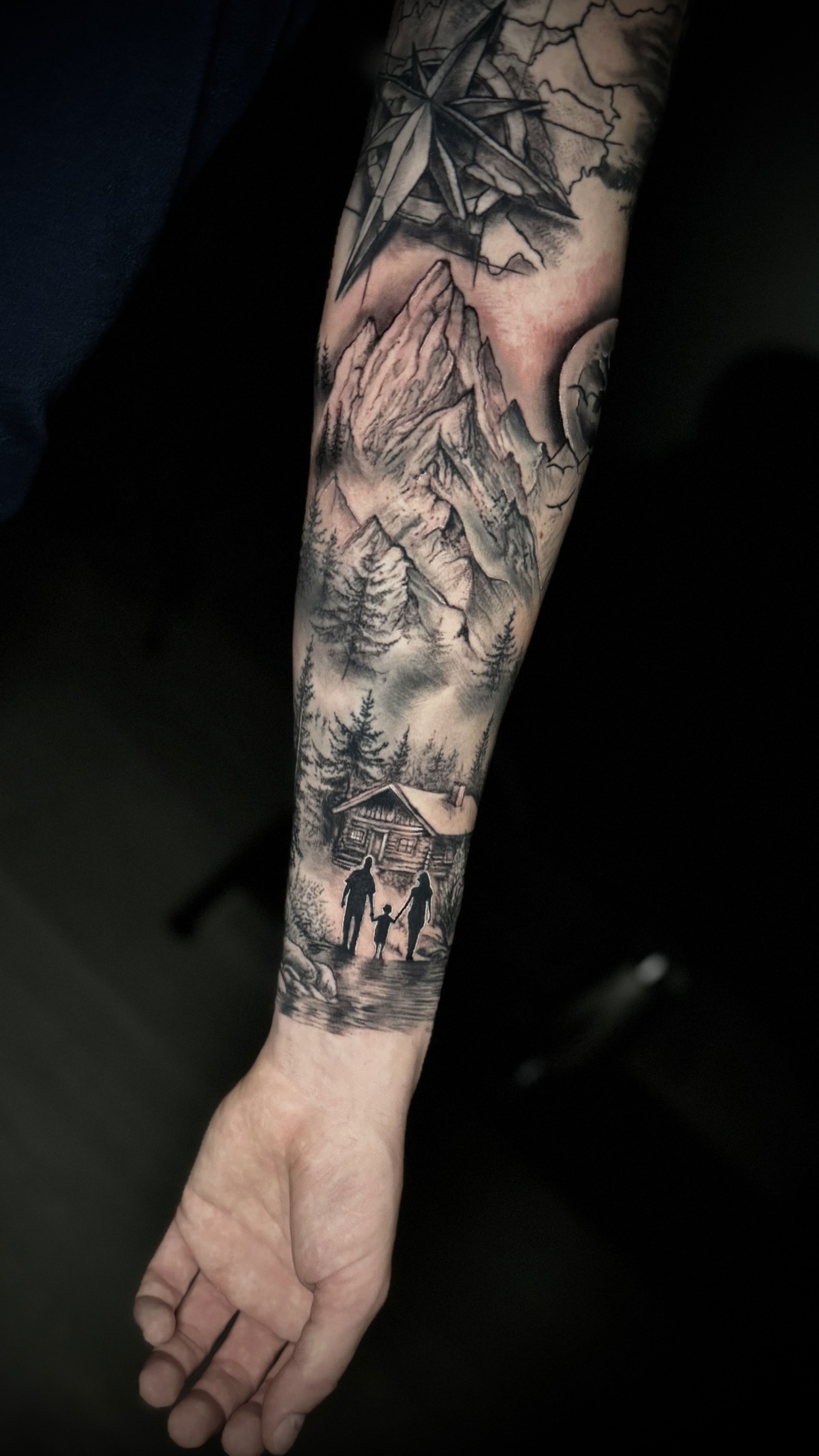 Tattoo auf dem Unterarm mit Berglandschaft, Wald, Mond und einem Haus, sowie einer Familie, die Hand in Hand am Fluss entlang geht.