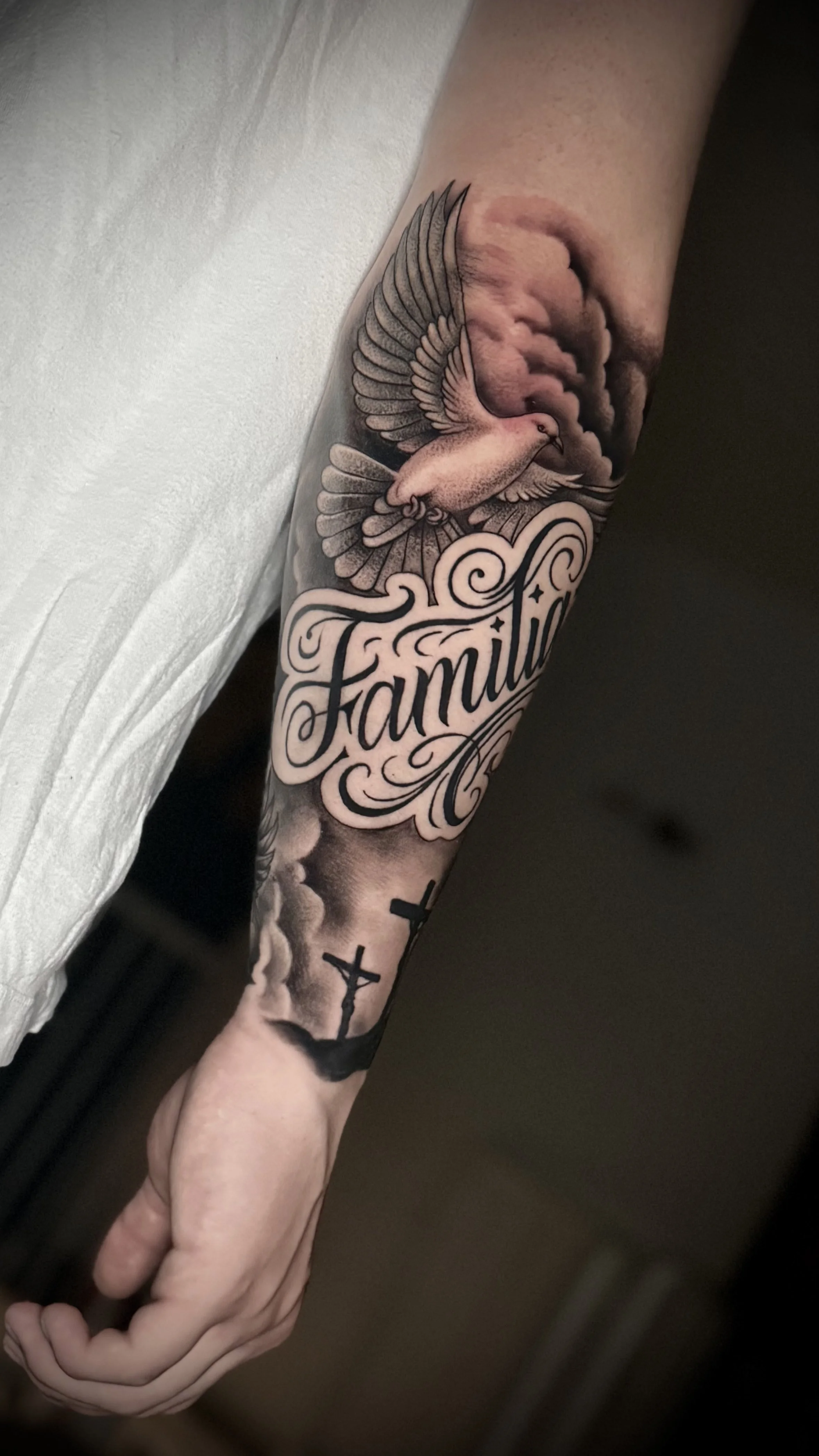 Realistische Black-and-Grey Tätowierung mit Taube, Schriftzug „Familie“ und Kreuzen auf dem Unterarm.