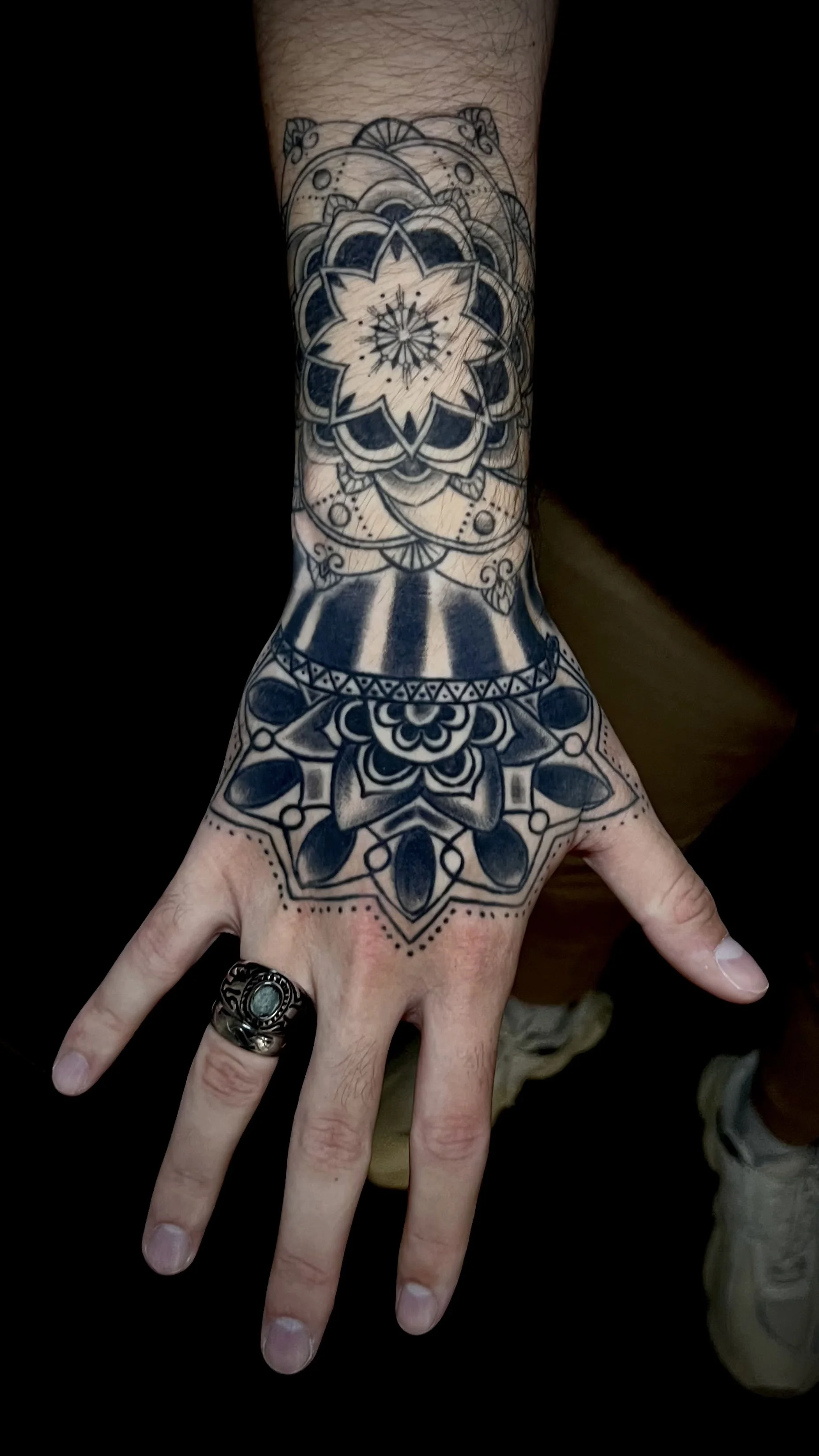Hände mit schwarzer Mandala-Tätowierung am Handgelenk und auf dem Handrücken, getragen mit einem Silberring mit grünem Stein.