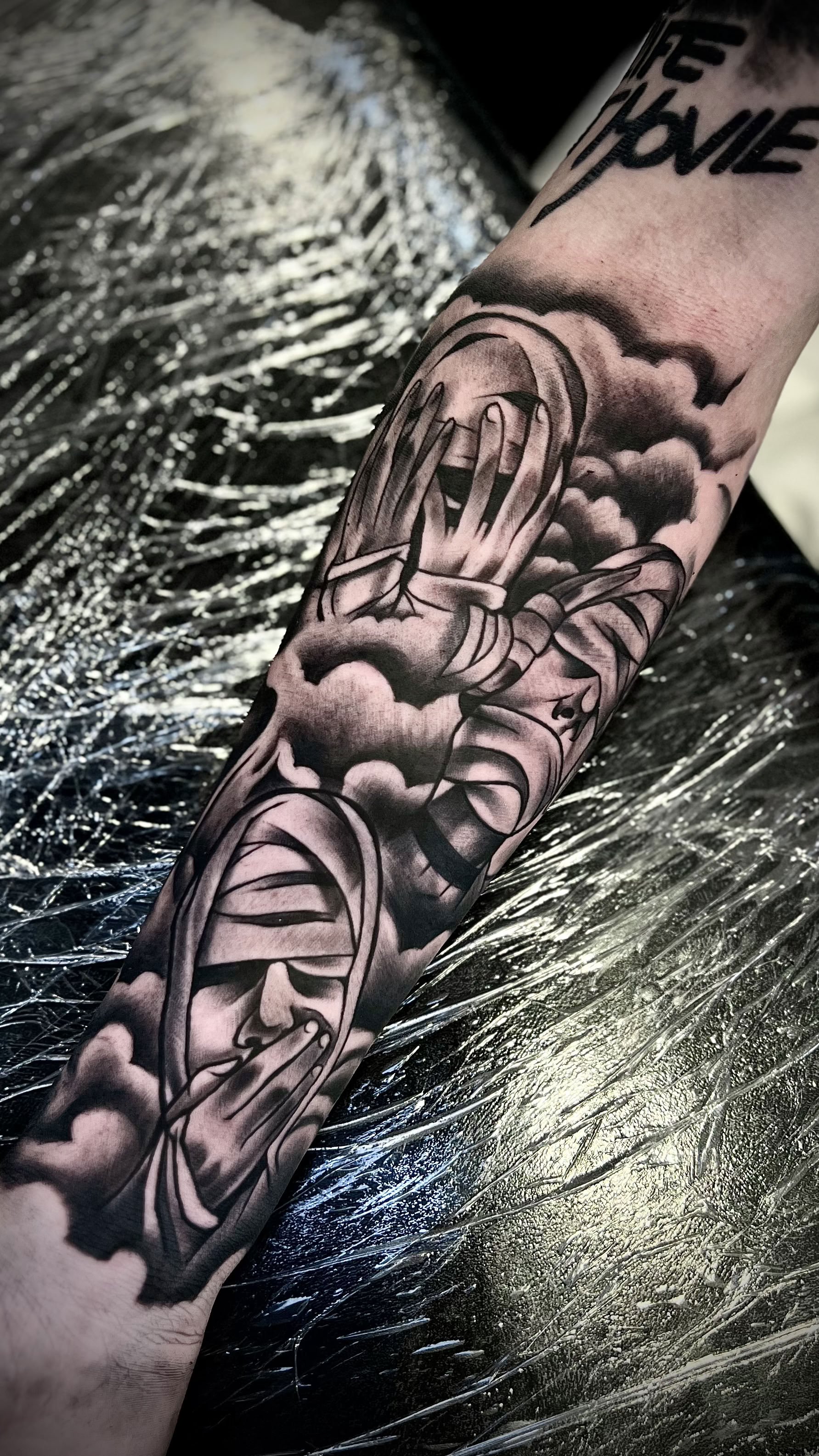 Schwarz-weiße Tätowierung eines Comics oder Illustrations auf einem Arm, zeigt eine Person mit geöffnetem Mund, Wolken und andere Designs.