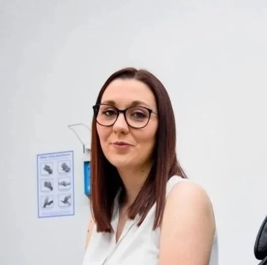 Eine Frau mit Brille sitzt im medizinischen Umfeld, vermutlich auf einem Untersuchungstisch.